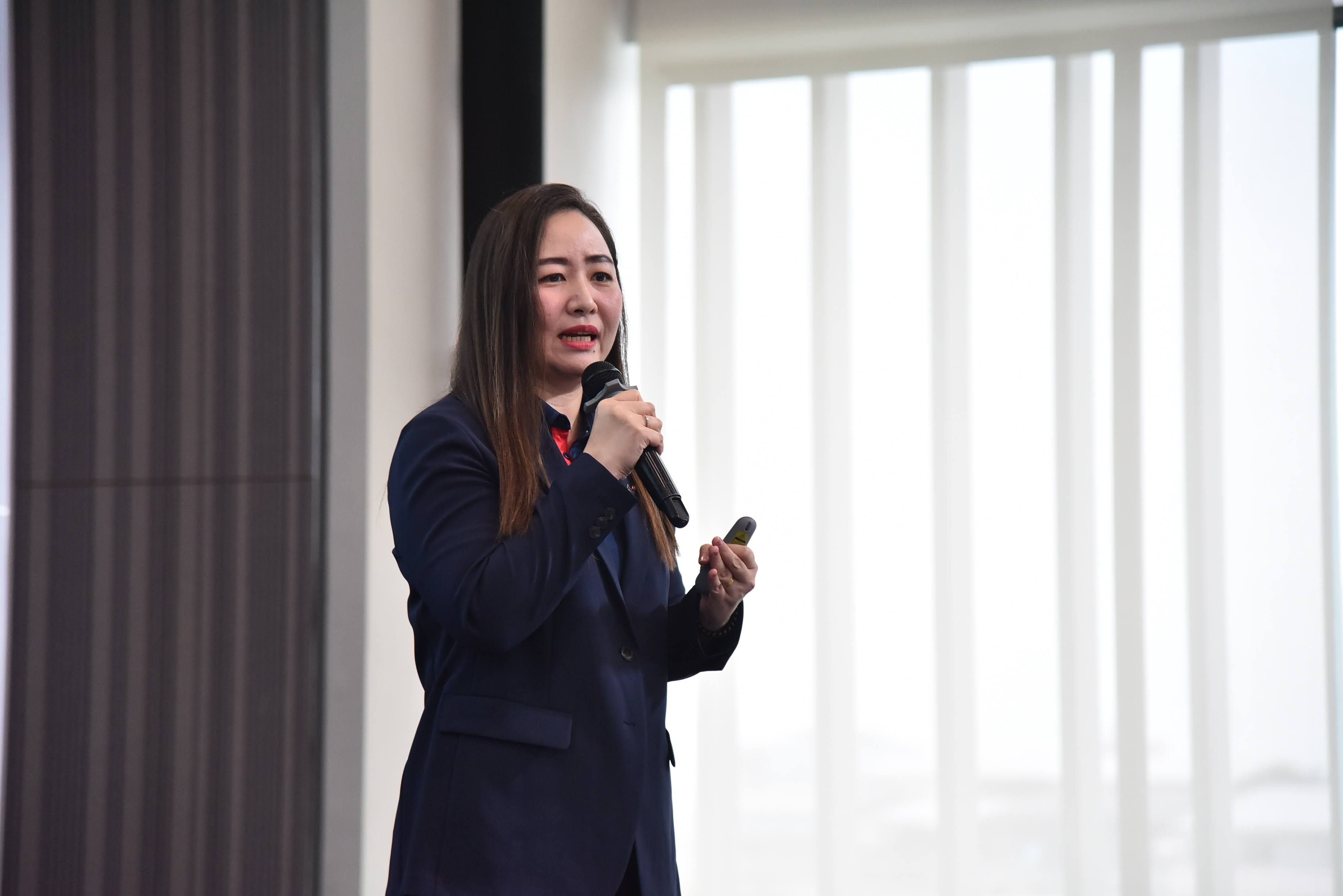 A woman in a dark suit speaks into a microphone, gesturing during a presentation.