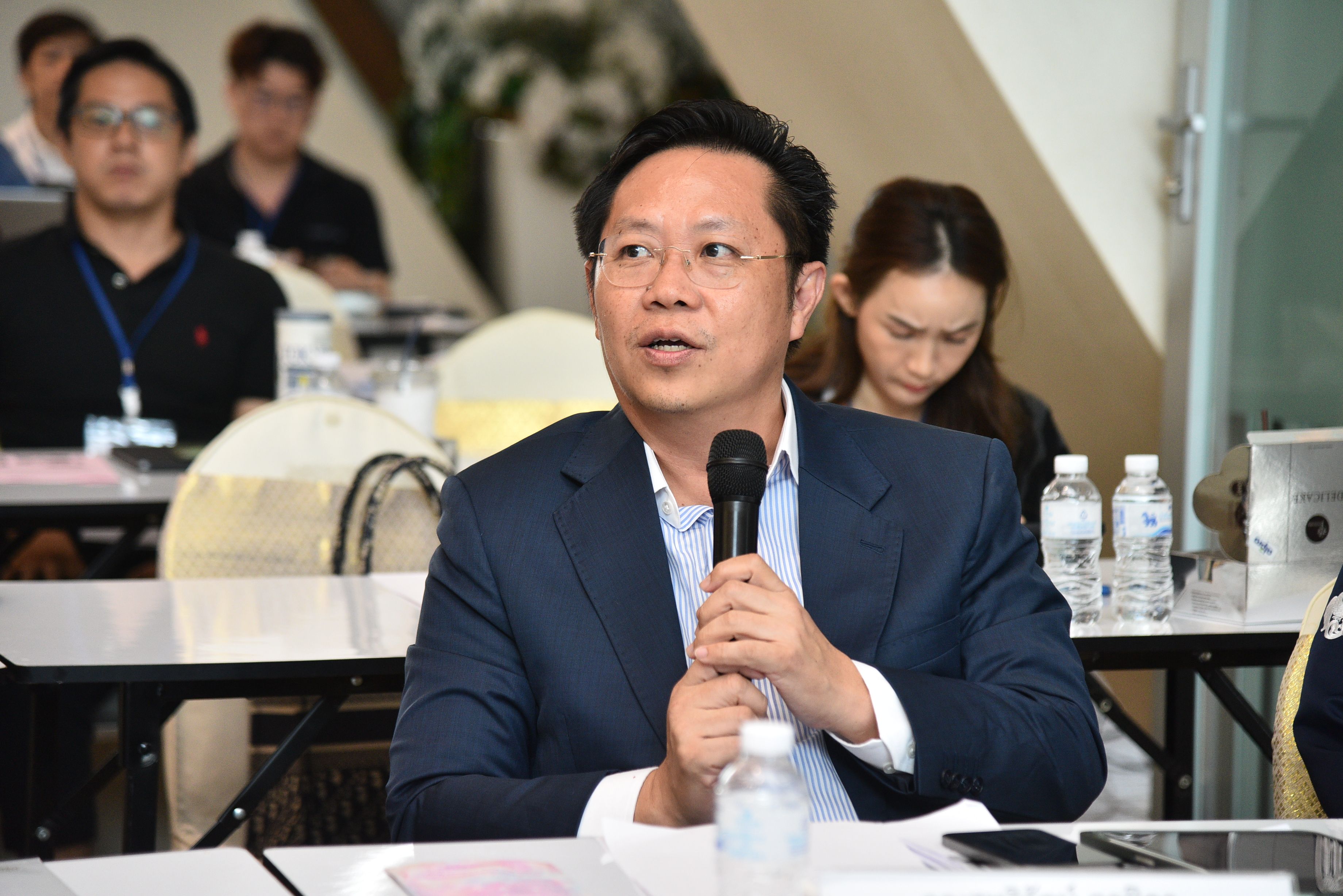 Professional Asian man in a dark suit speaks into a microphone at a conference table.