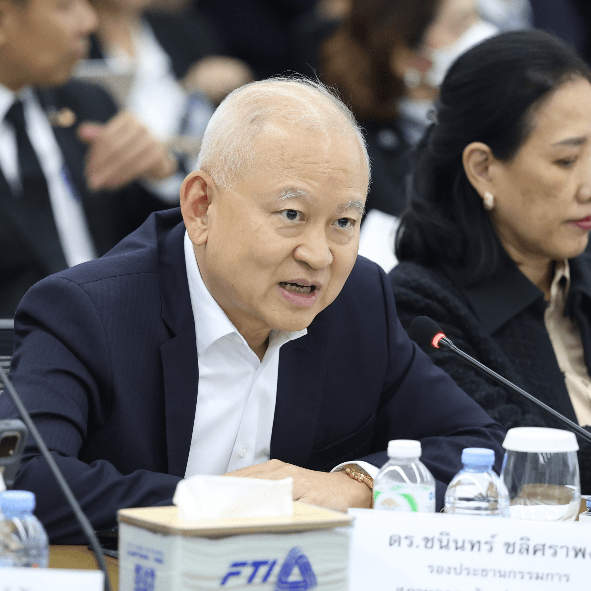 An elderly Asian man in a dark suit speaks animatedly at a business meeting.