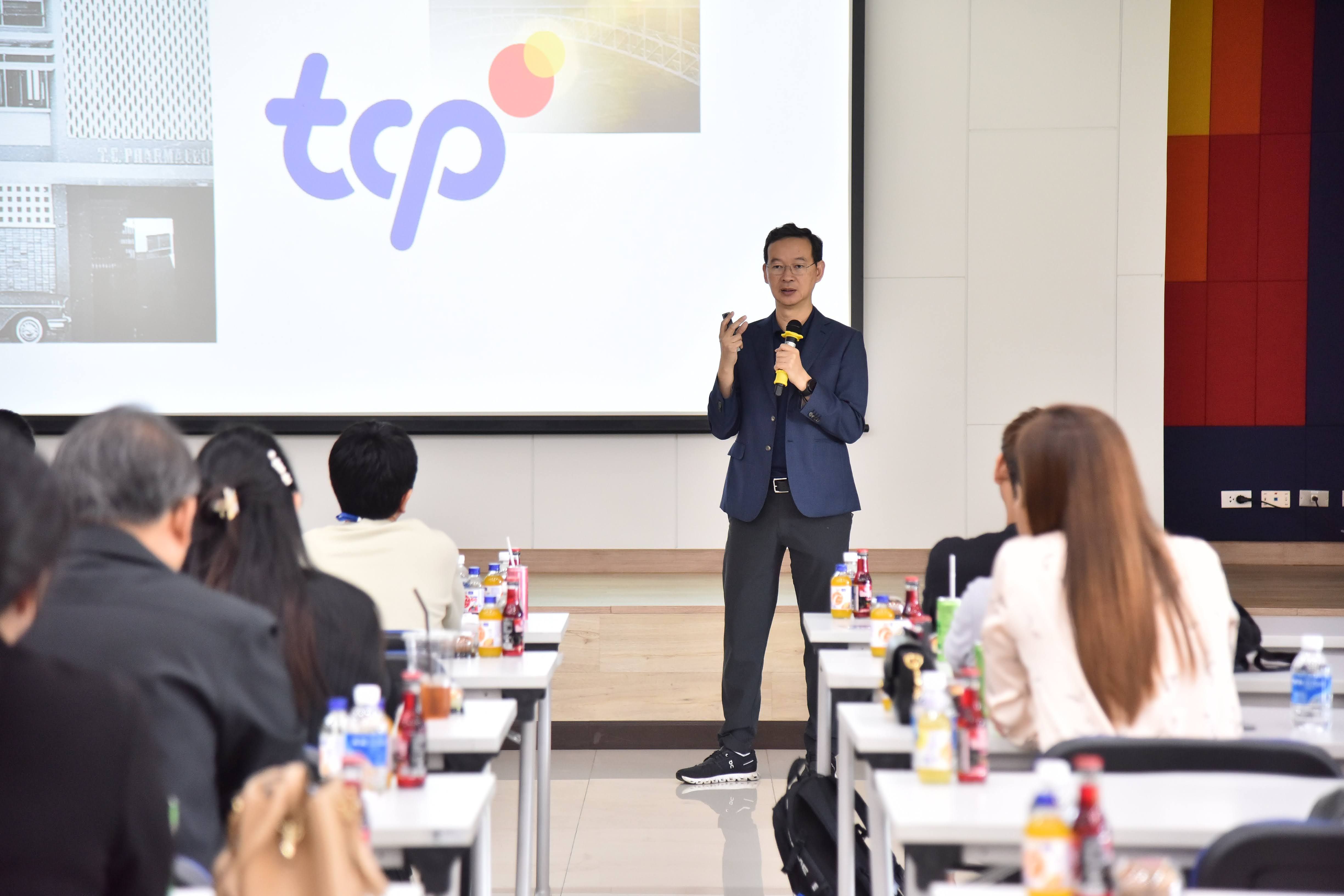 Man speaking into microphone in front of projection screen with 'tcp' logo.