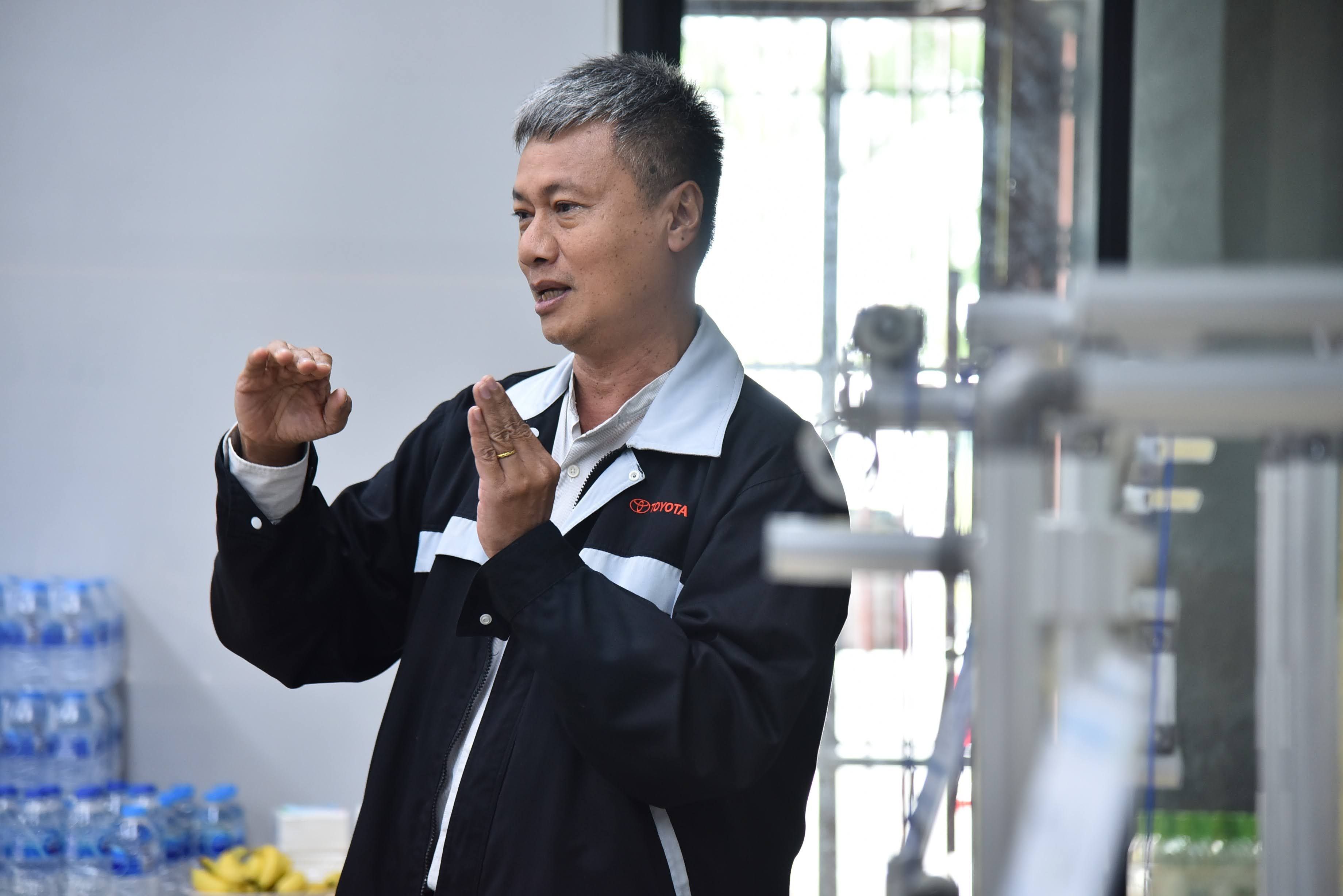 Middle-aged Asian man in a black jacket speaking and gesturing with both hands indoors.