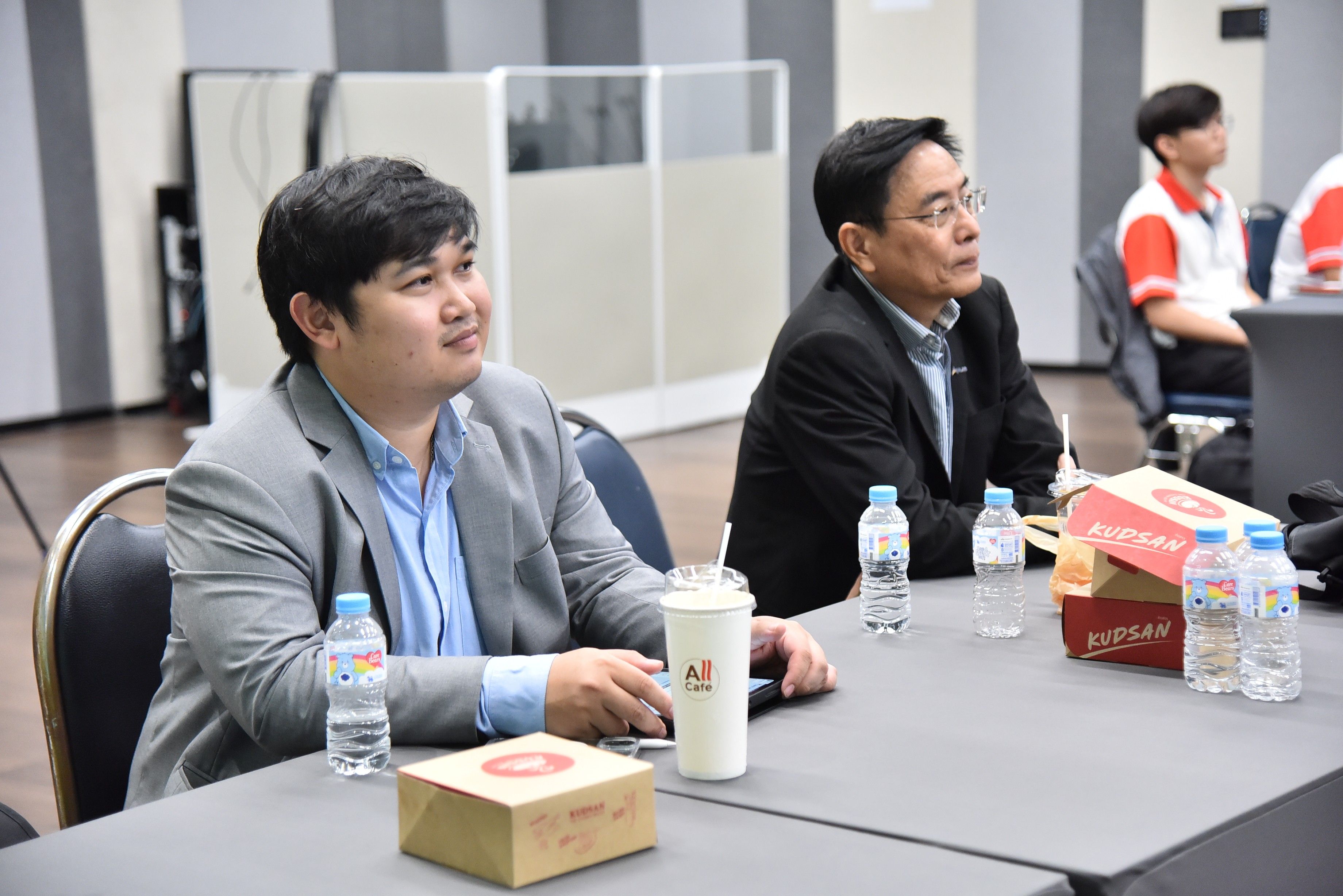 Two men seated at a table, attentively looking forward with drinks and food.