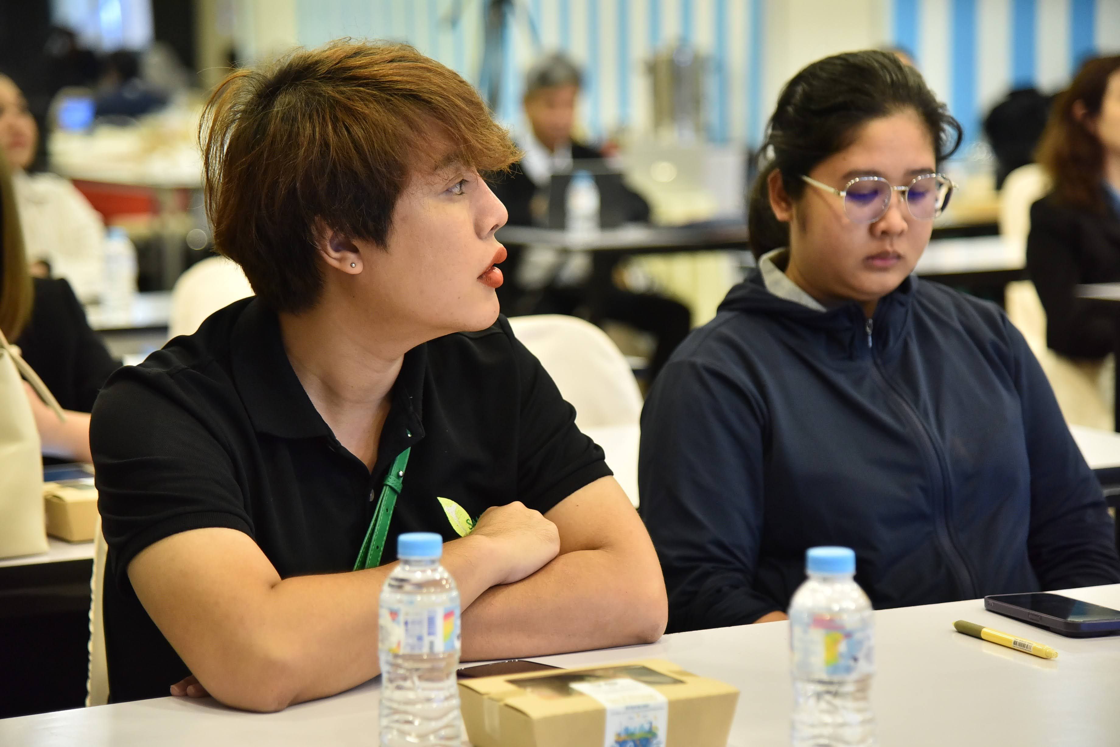 Two conference attendees, a man with brown hair and a woman in glasses, sit at a table.