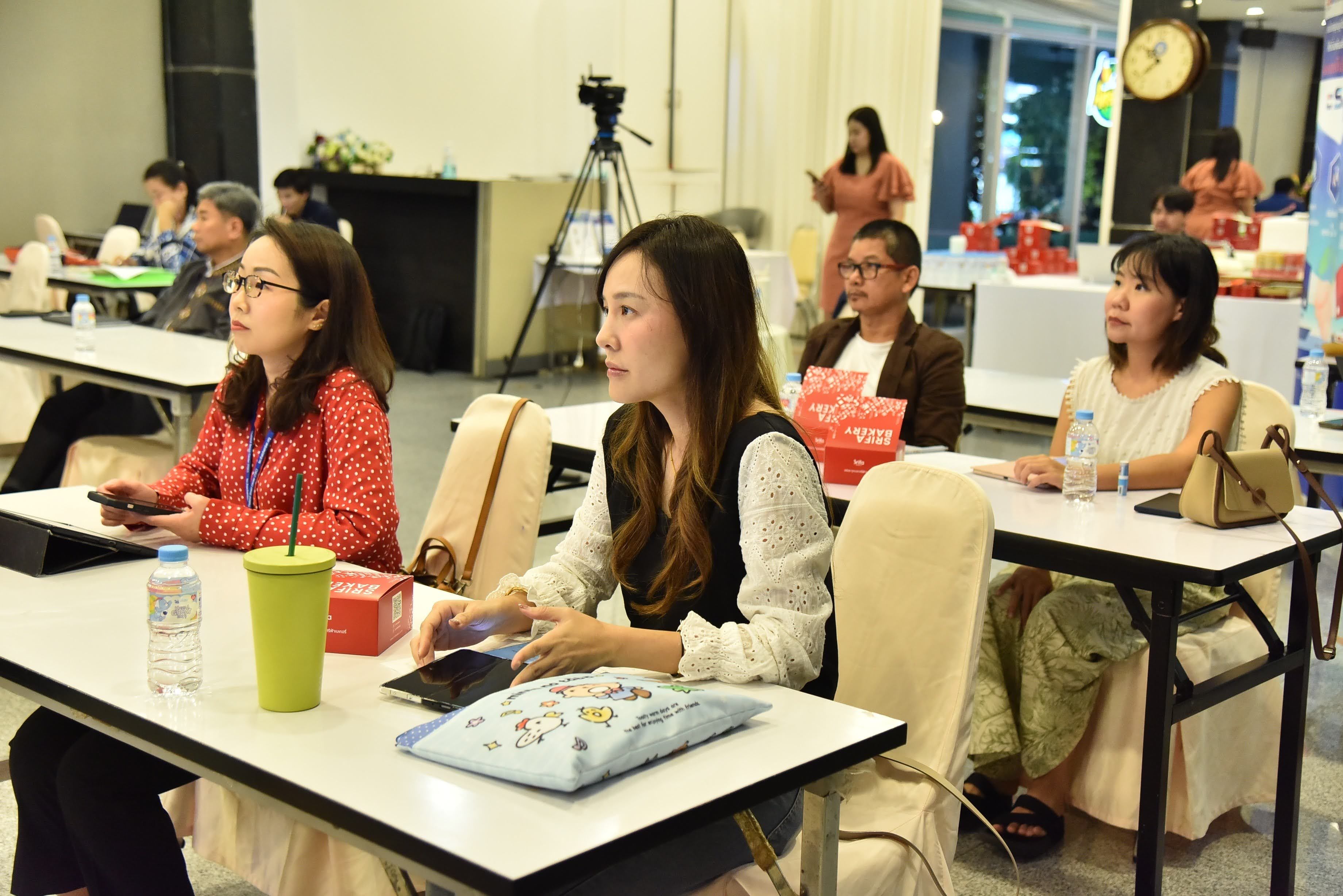Attendees listen attentively at a seminar, seated at tables with drinks and notebooks.
