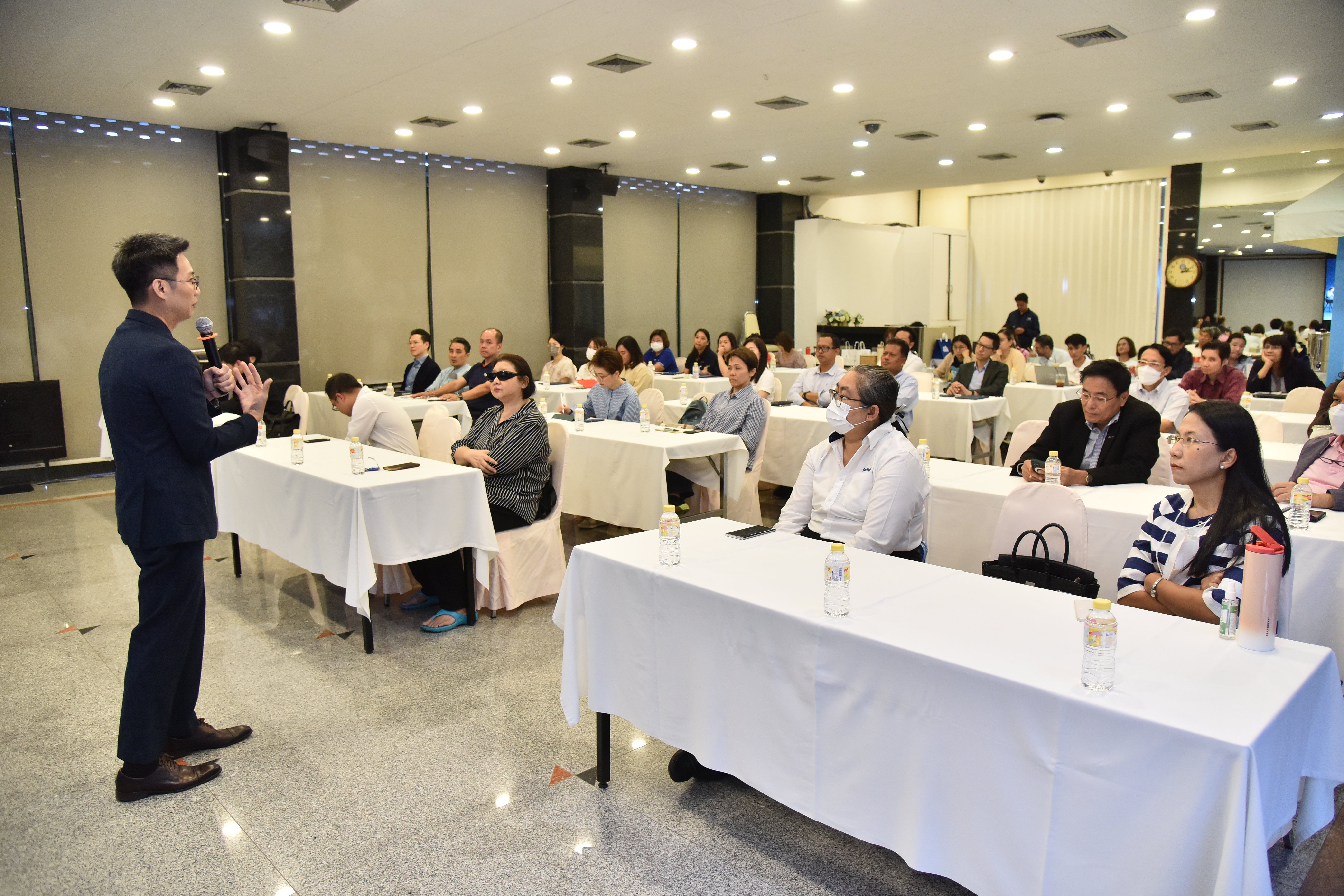 A man in a suit presents to an audience seated at tables in a conference room.