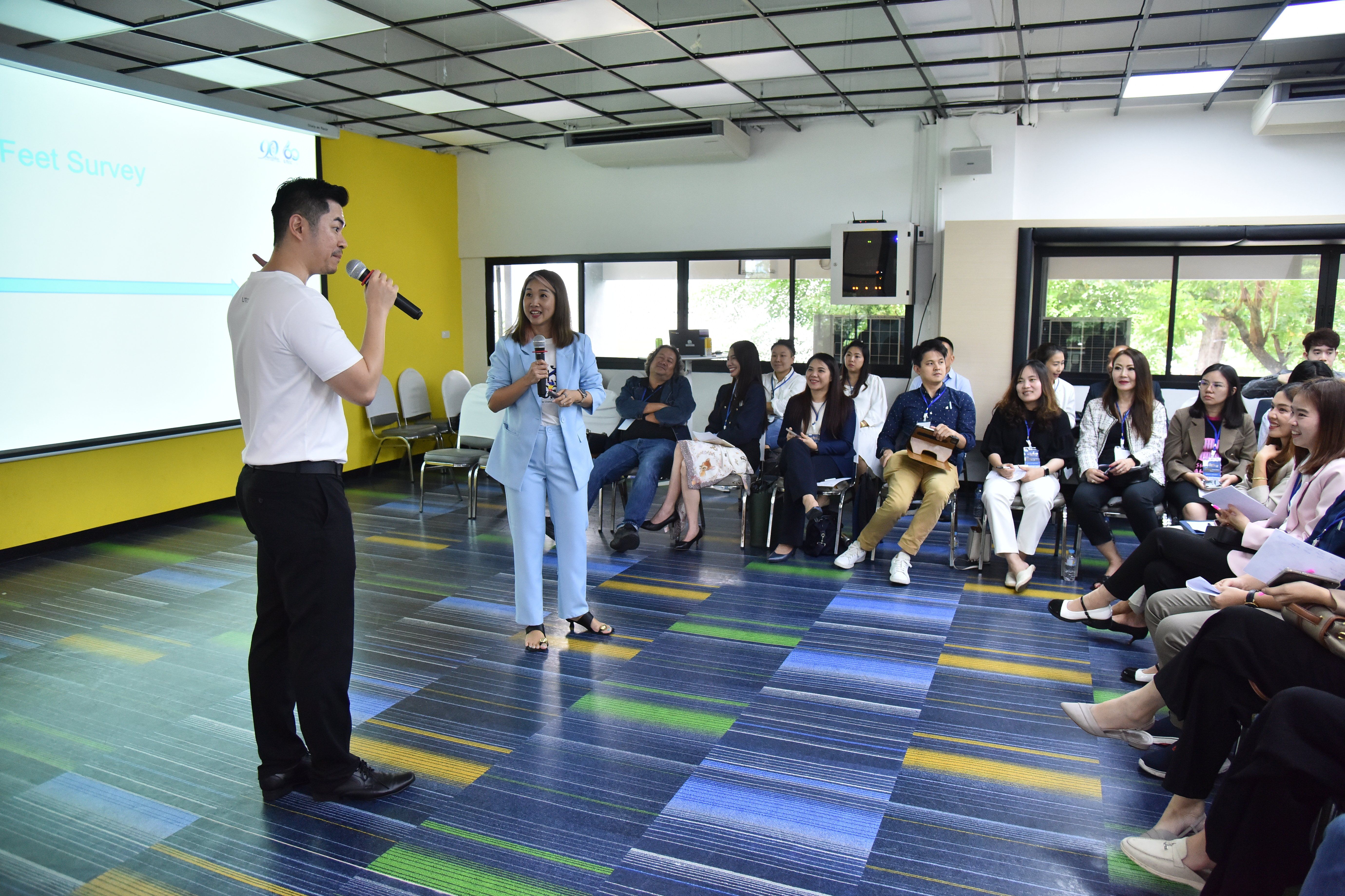 Man speaks into microphone next to woman, addressing a seated audience in a bright room.