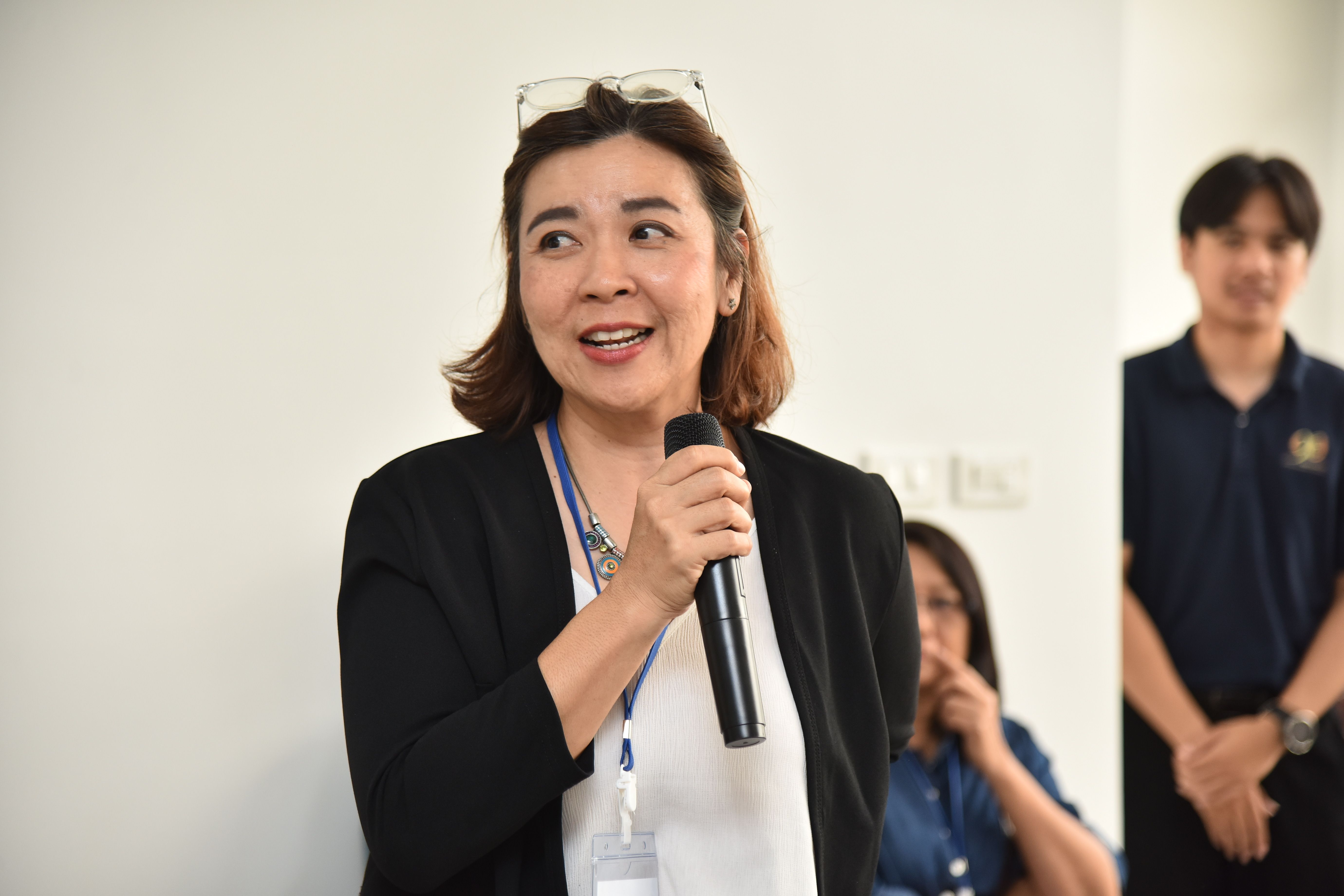 Middle-aged Asian woman speaking into a microphone, wearing glasses and a black blazer.