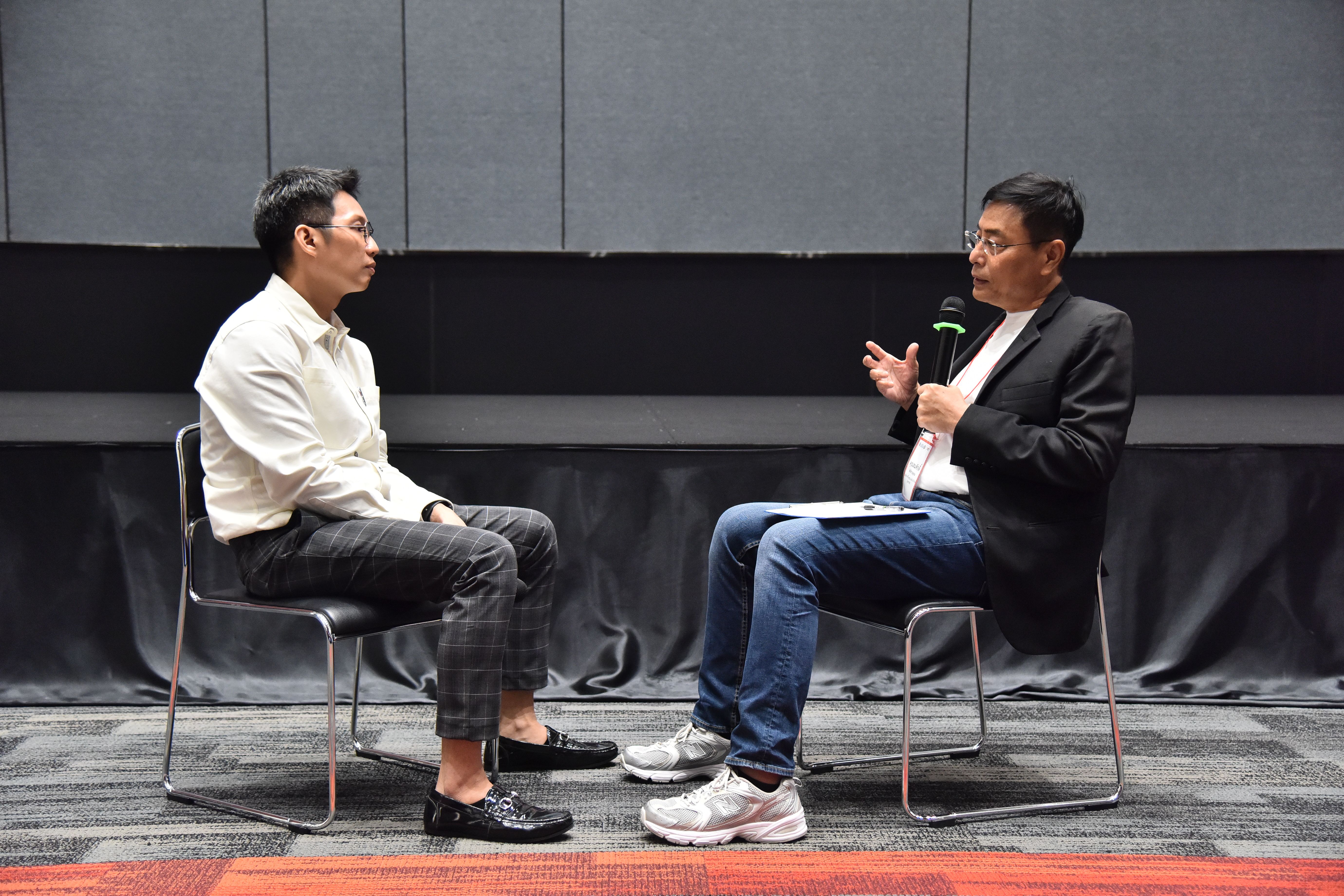 Two men sit facing each other, one holding a microphone during an interview.