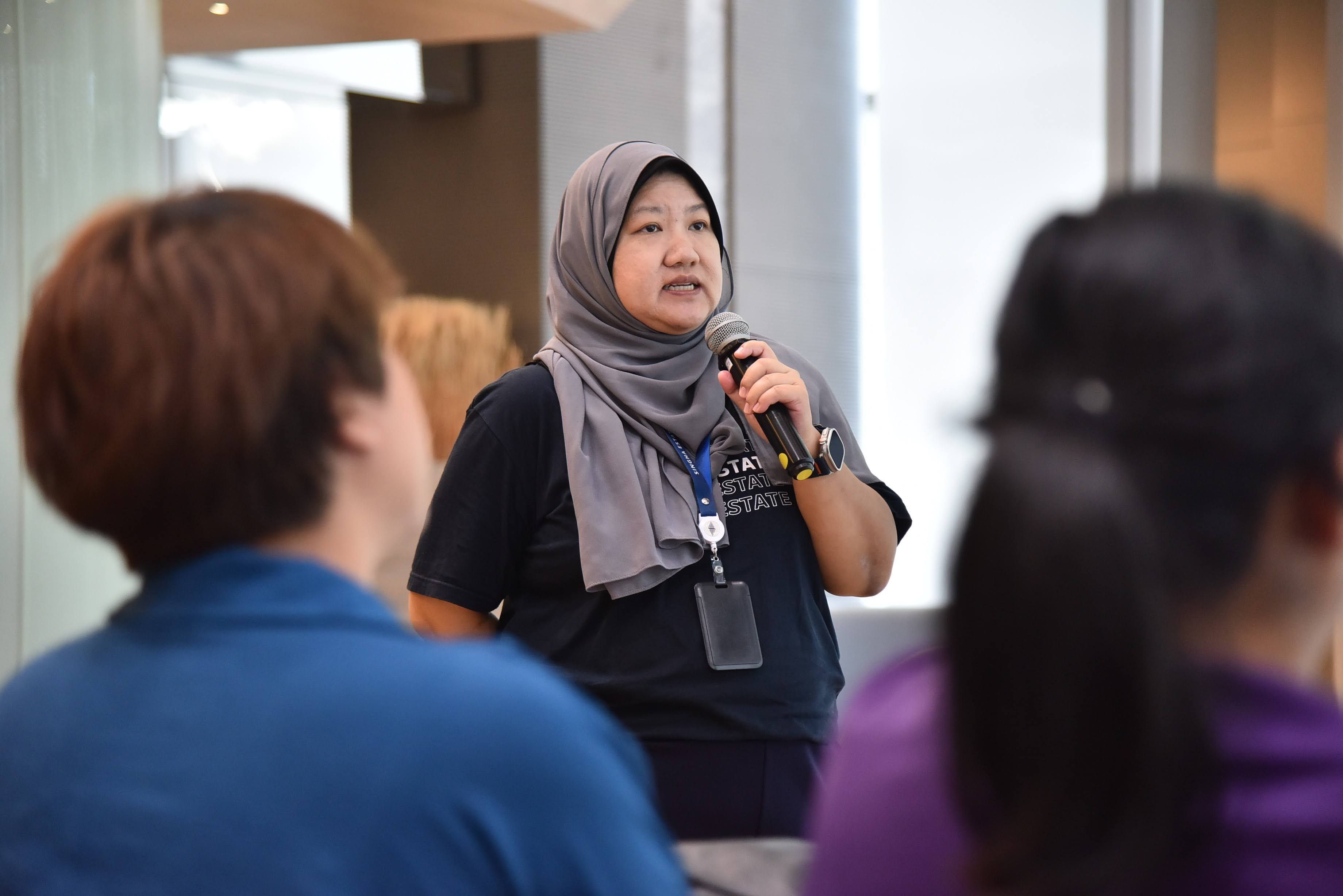 Woman in grey hijab speaks into microphone addressing an audience during a presentation.