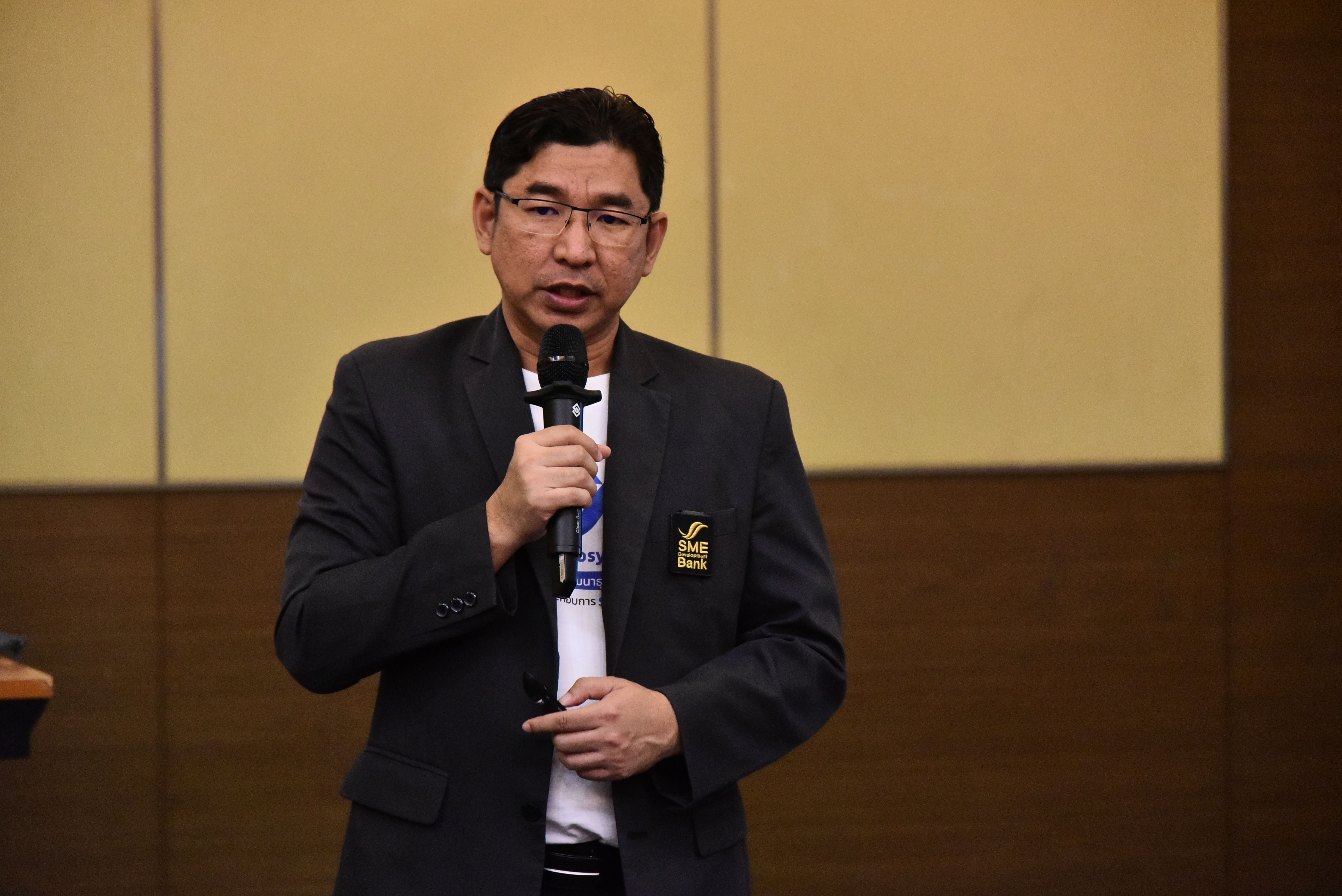 Man in glasses and suit jacket speaking into a microphone at a professional presentation.