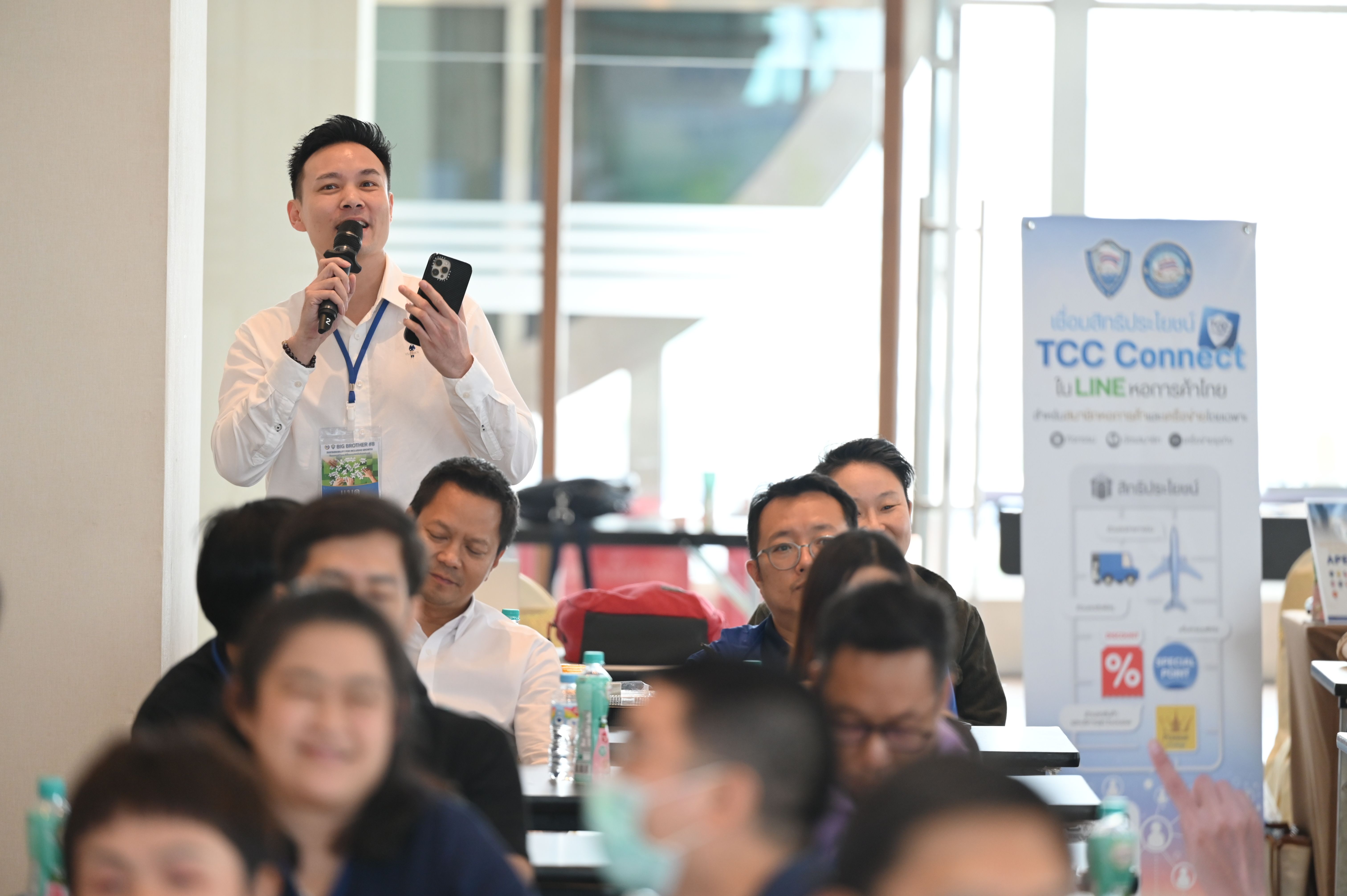 Man in white shirt speaking into microphone at a professional event with an audience.