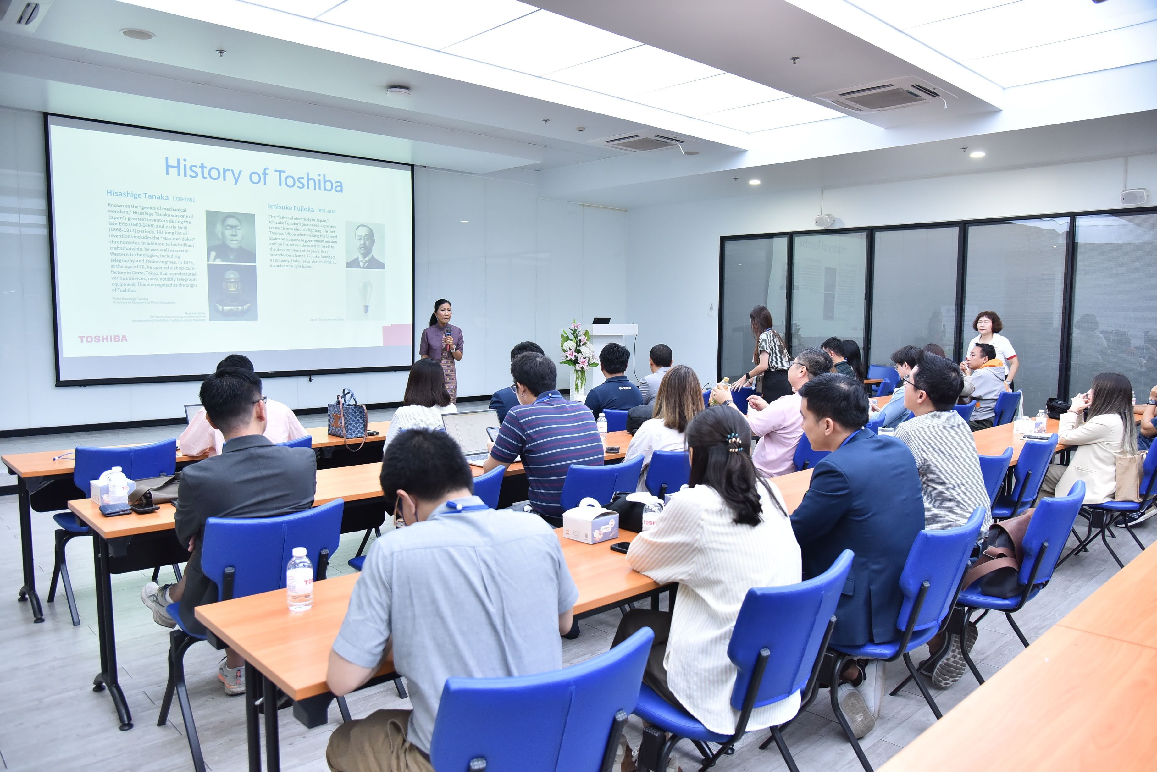 Audience attends a presentation in a brightly lit room with a speaker and projector showing slides.