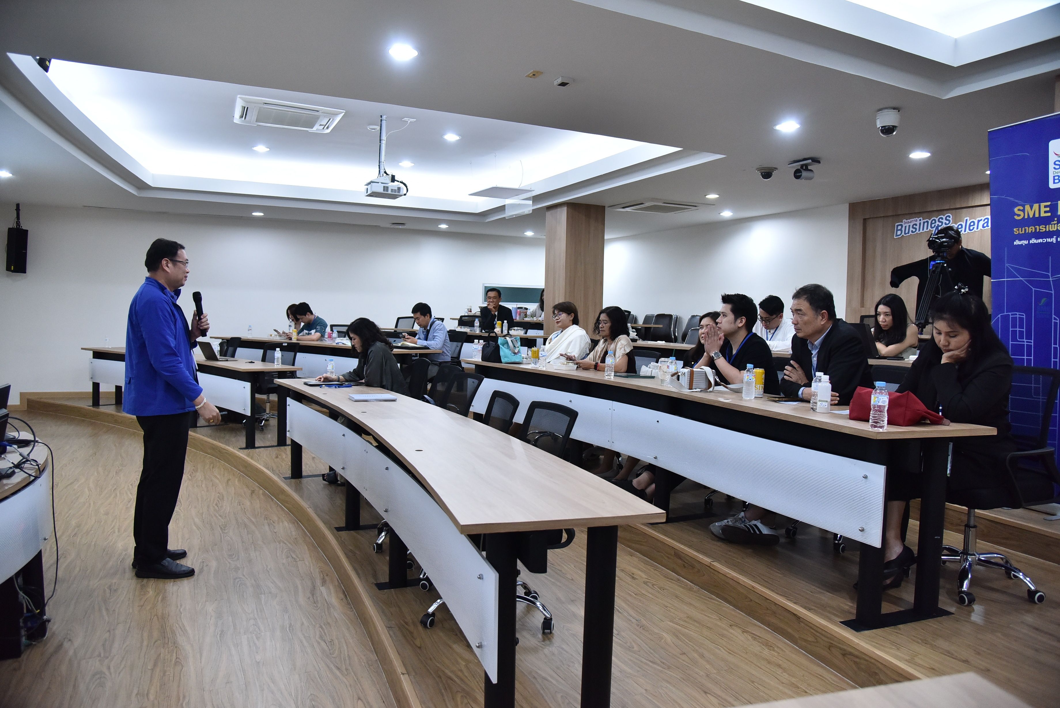 A man in a blue jacket speaks to an attentive audience in a modern classroom setting.