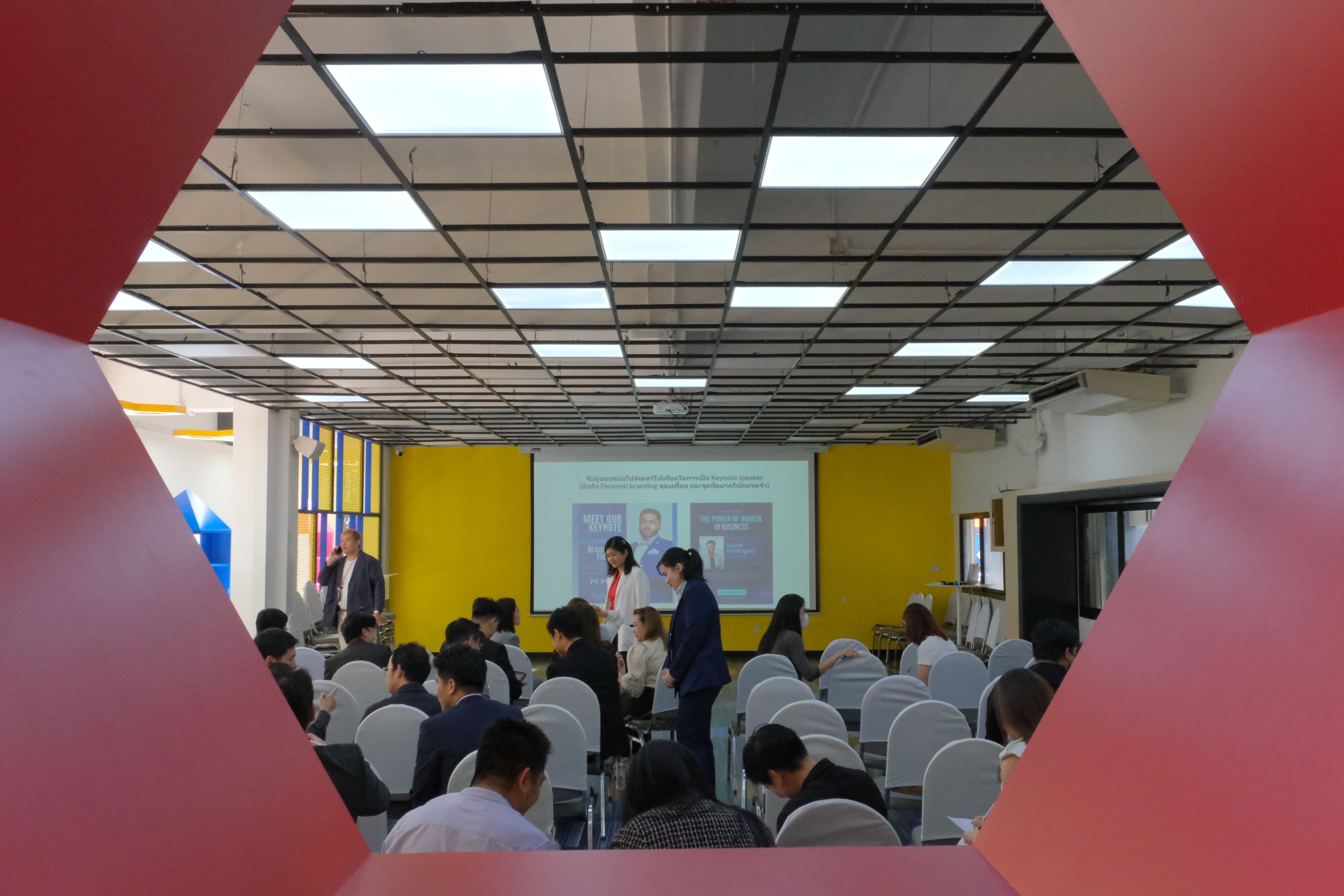 People attend a presentation in a modern conference room with a large screen and yellow wall.
