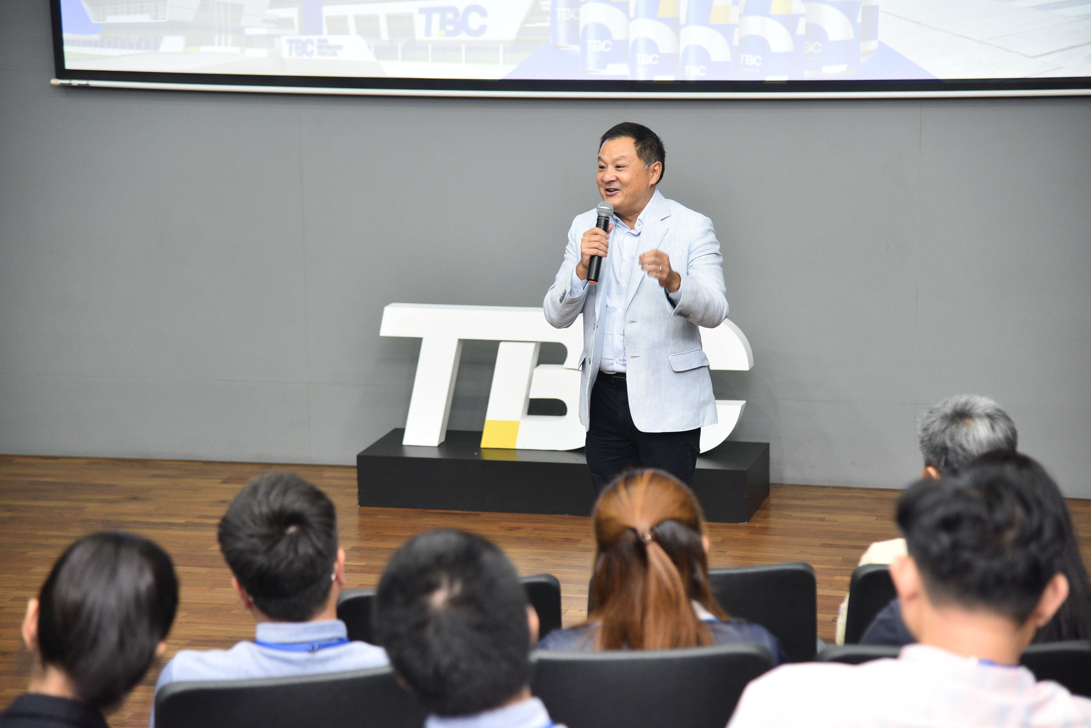 Man speaking on stage with microphone in front of a TBC logo to an audience.