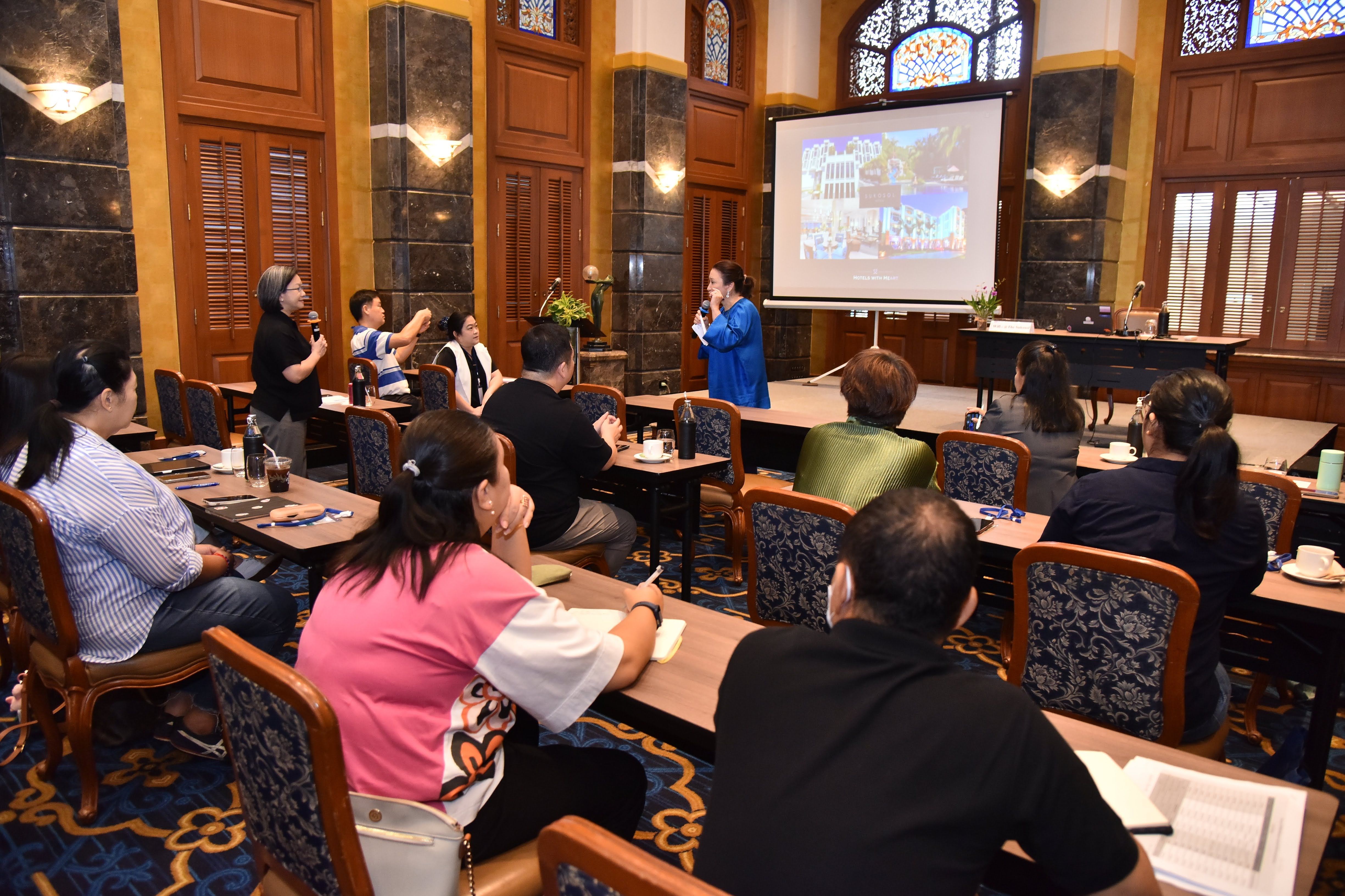 A woman presents to a seated audience in a warmly lit conference room with a projector.