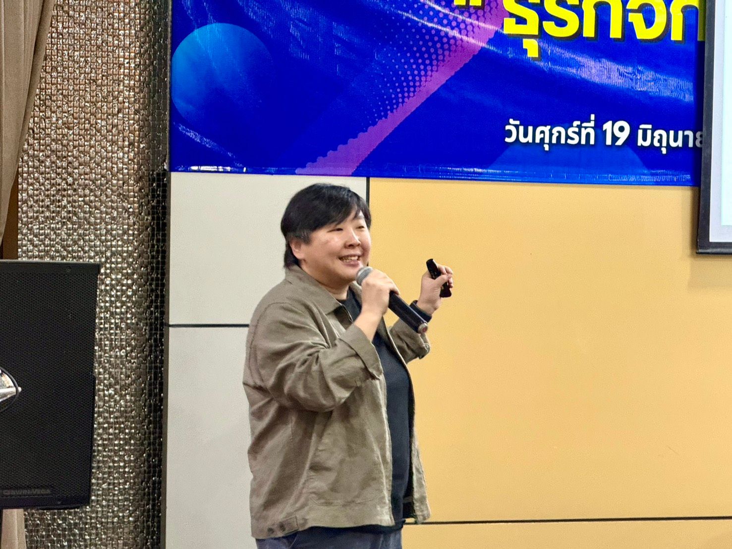 Woman in brown jacket speaking into a microphone, holding a remote.