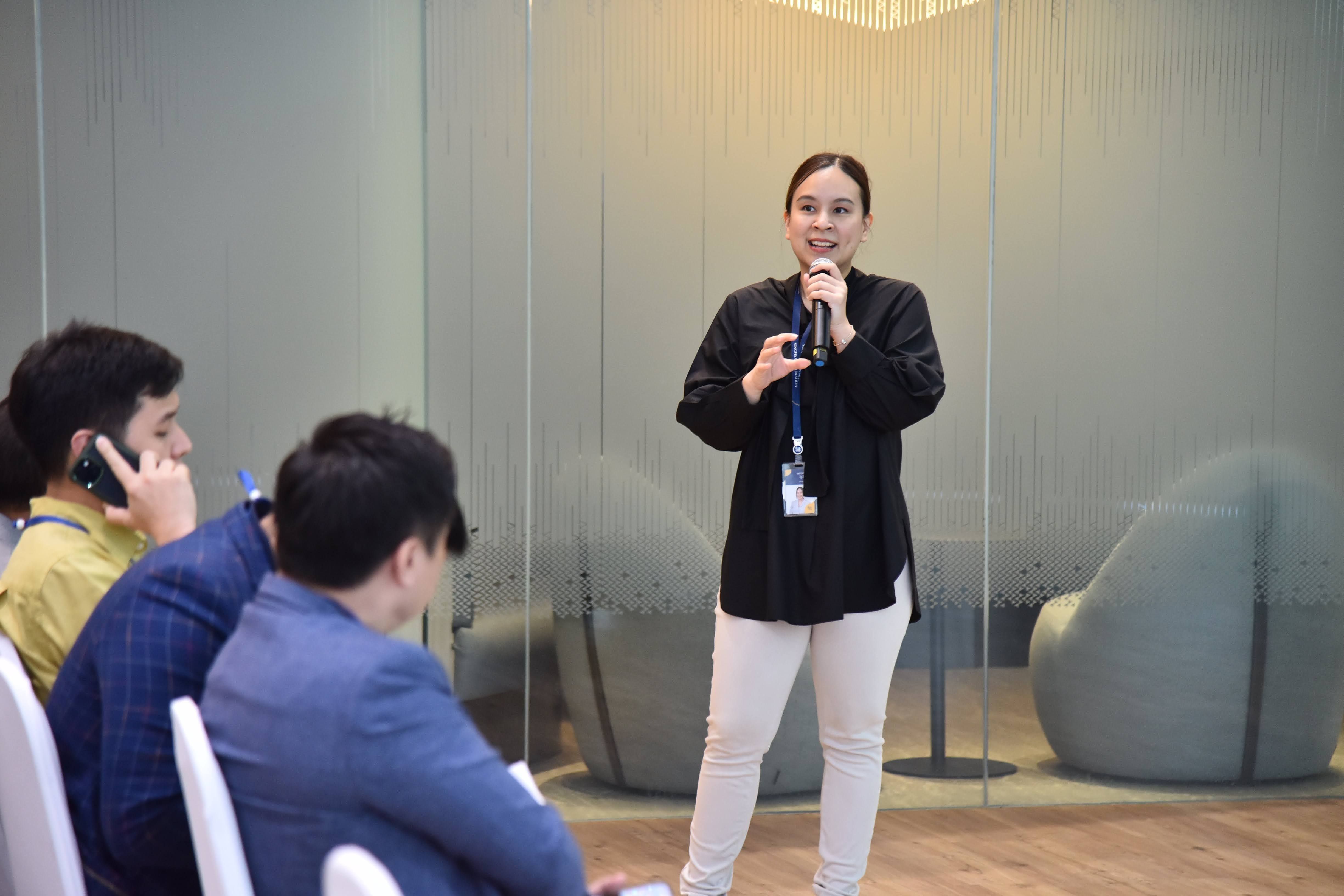 A woman stands confidently, speaking into a microphone to a seated audience in a modern room.