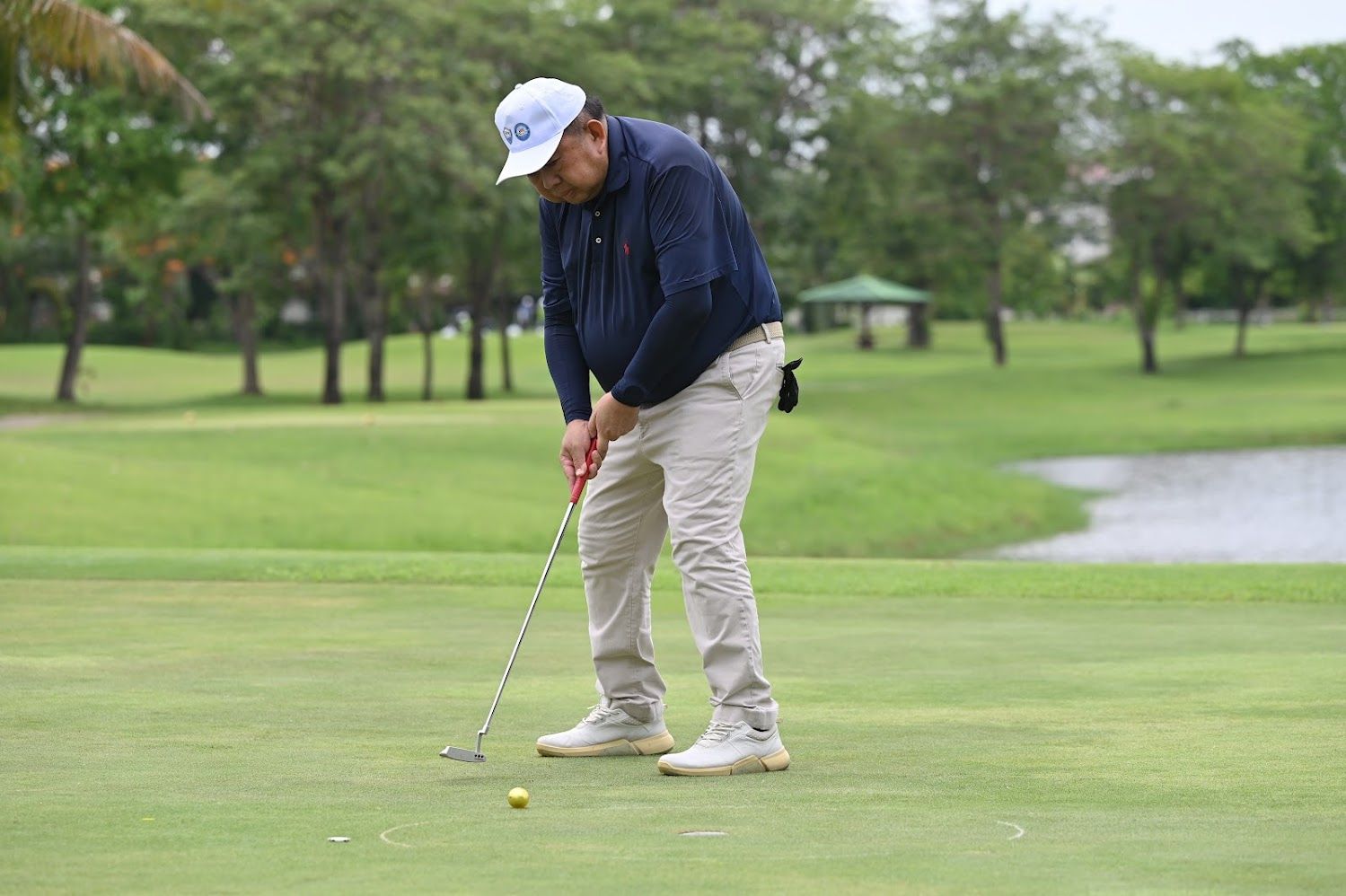 Man in blue jacket and white cap putting a yellow golf ball on a green course.
