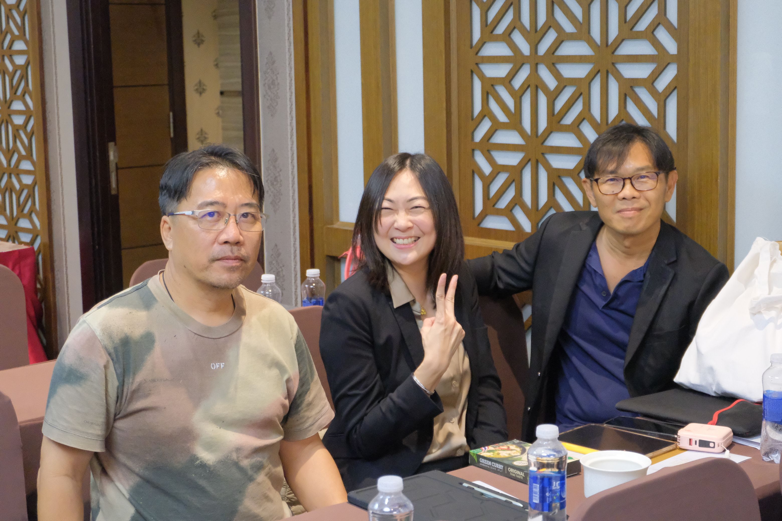 Three smiling people, two men and one woman, sitting at a table with a patterned wall.