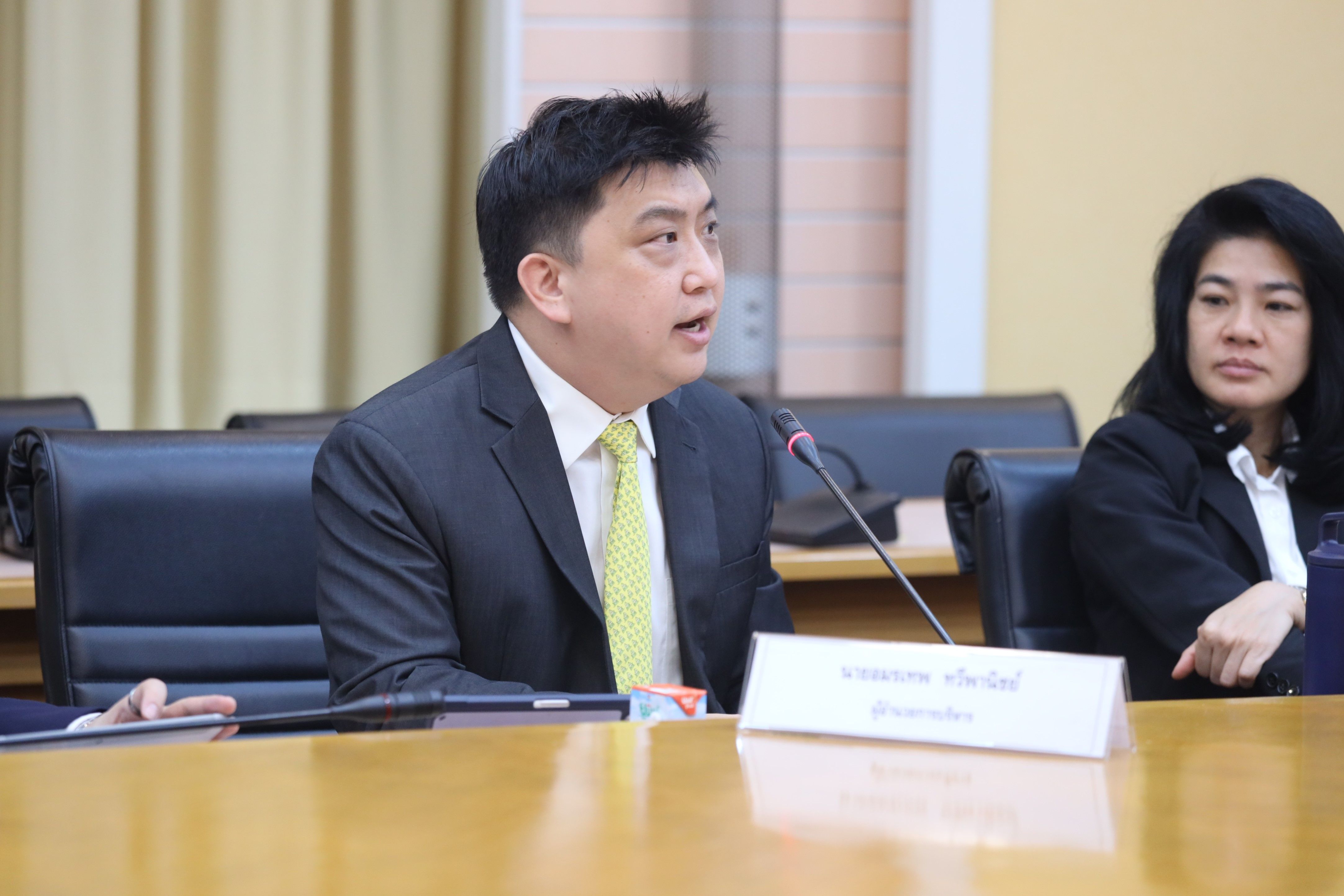 Man in a dark suit and light green tie speaking into a microphone at a conference table.