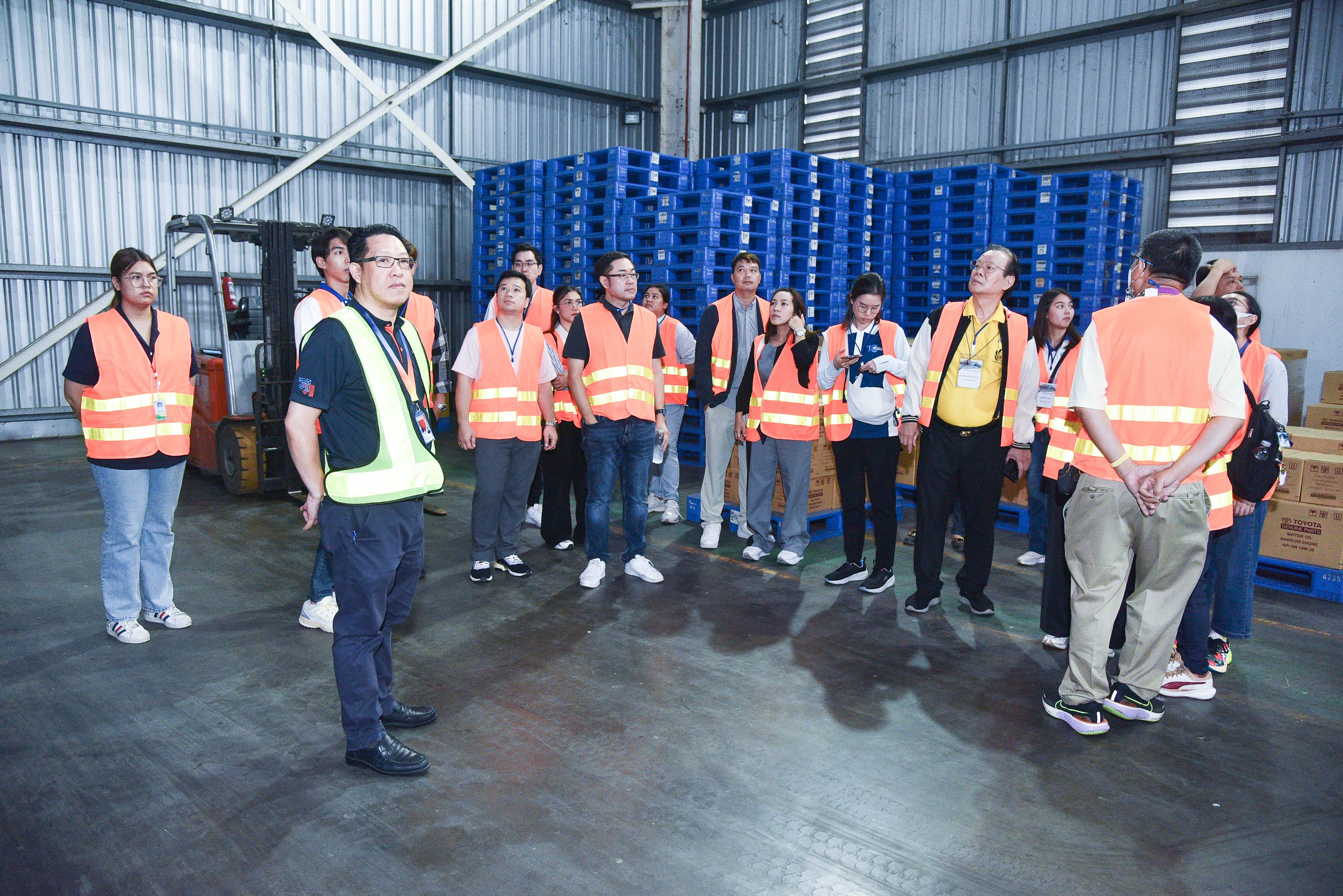 Group of people in orange safety vests inside a large warehouse with blue pallets.