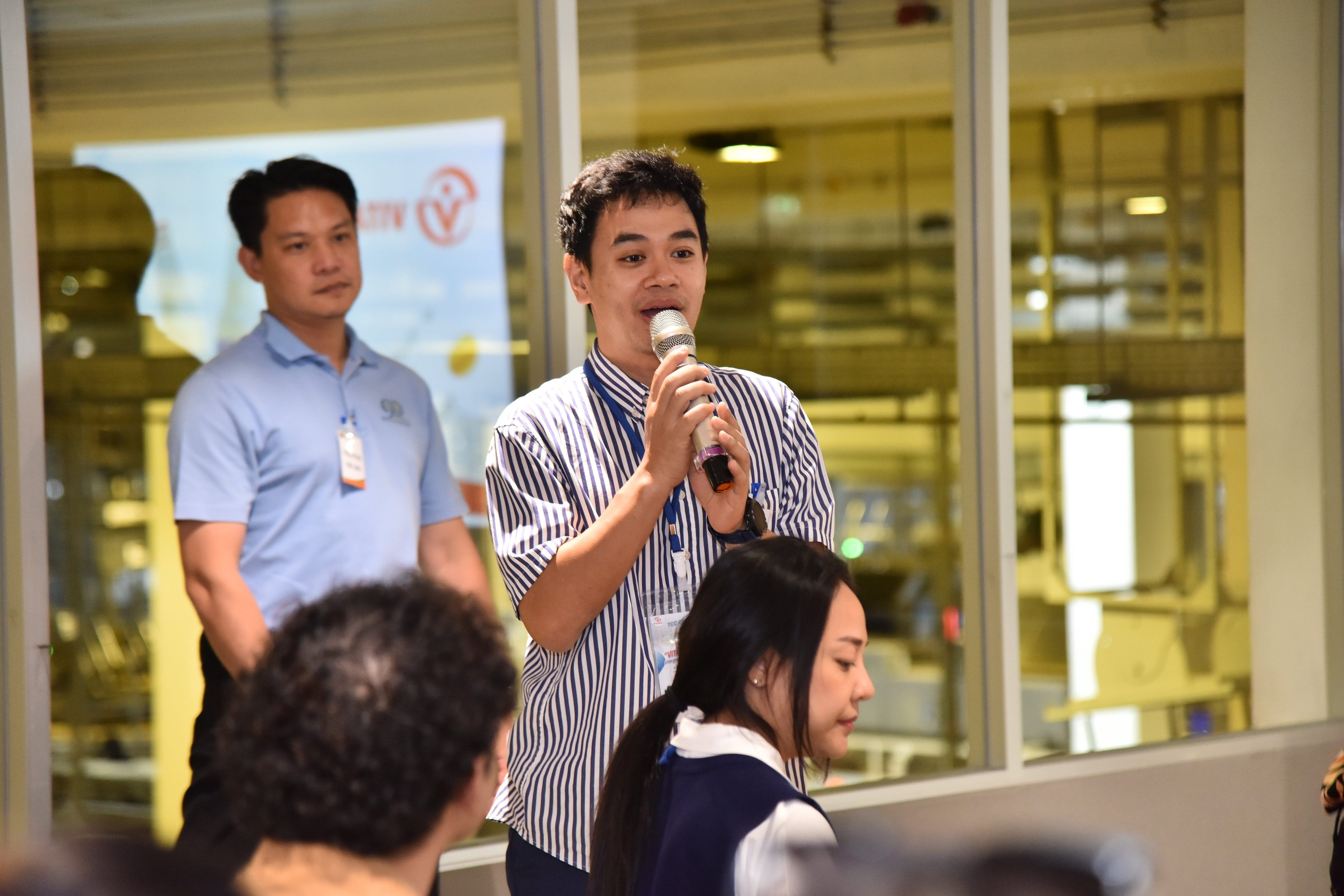 Man in striped shirt speaks into a microphone at an indoor event with a colleague.