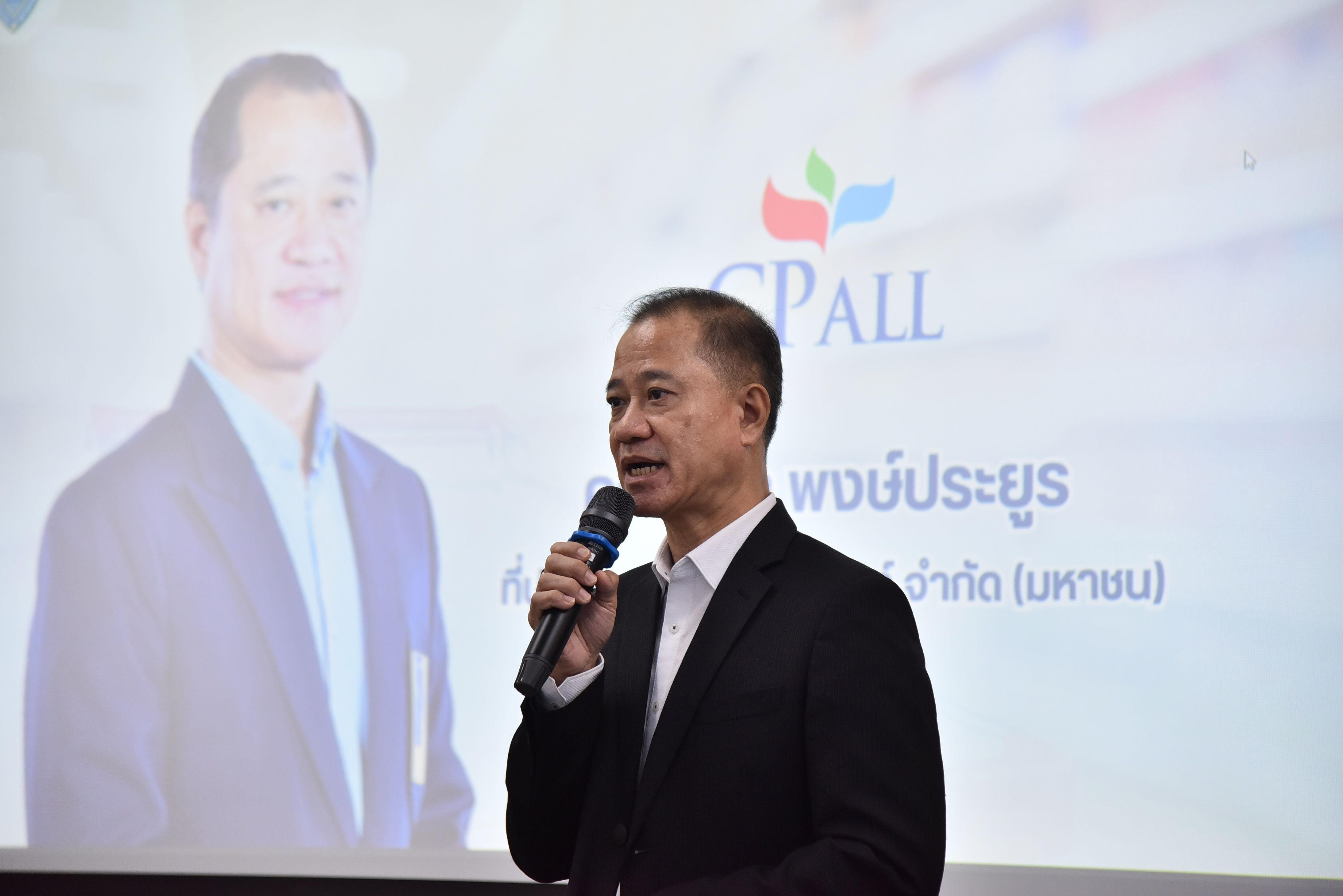 Man in dark suit speaks into microphone, CPALL logo visible on presentation screen.