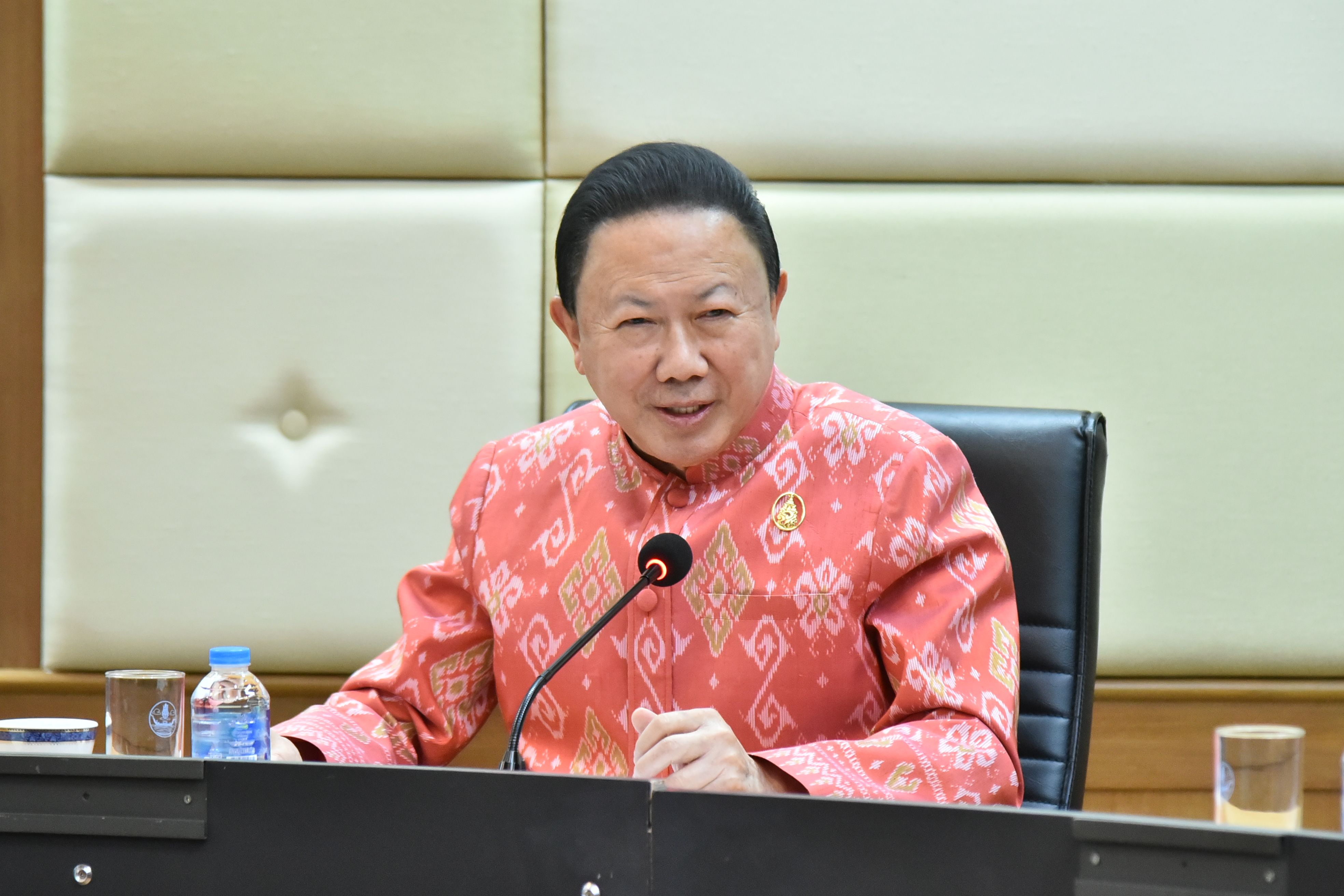 Older Asian man in traditional red patterned shirt speaking into a microphone at a table.