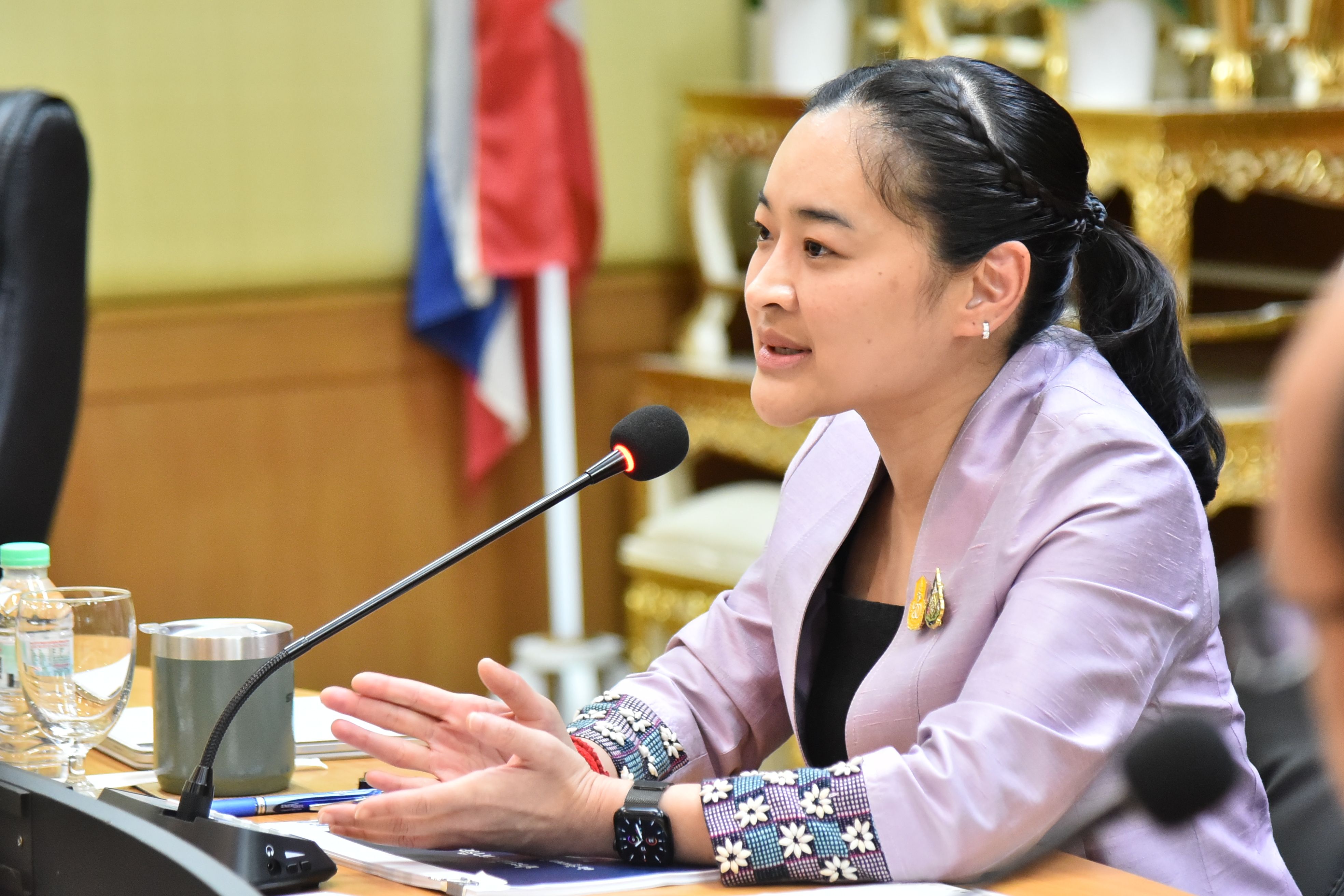 Woman in a lavender jacket speaking into a microphone during a formal meeting.