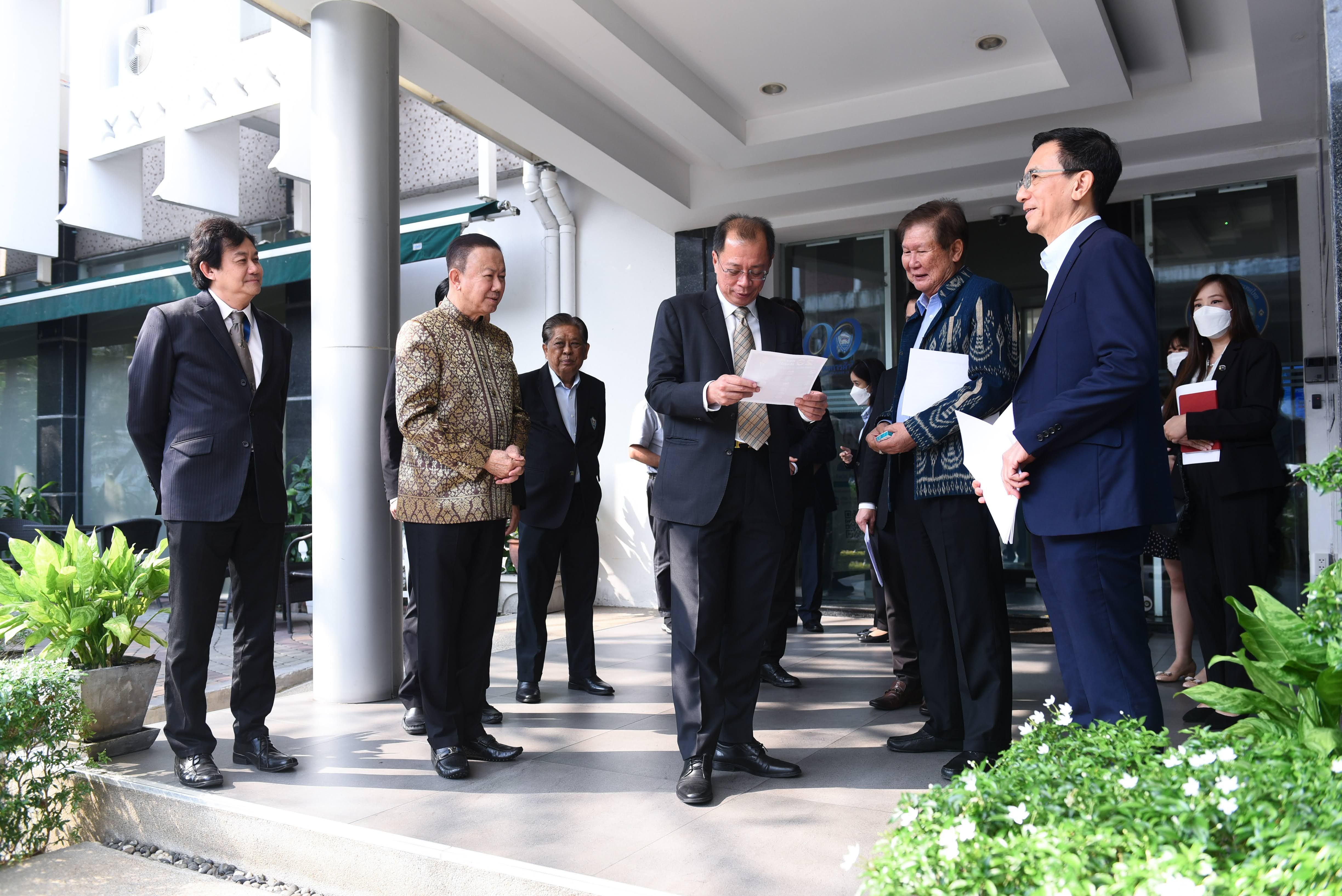 A group of delegates in formal and traditional attire meet outdoors, one reading a document.