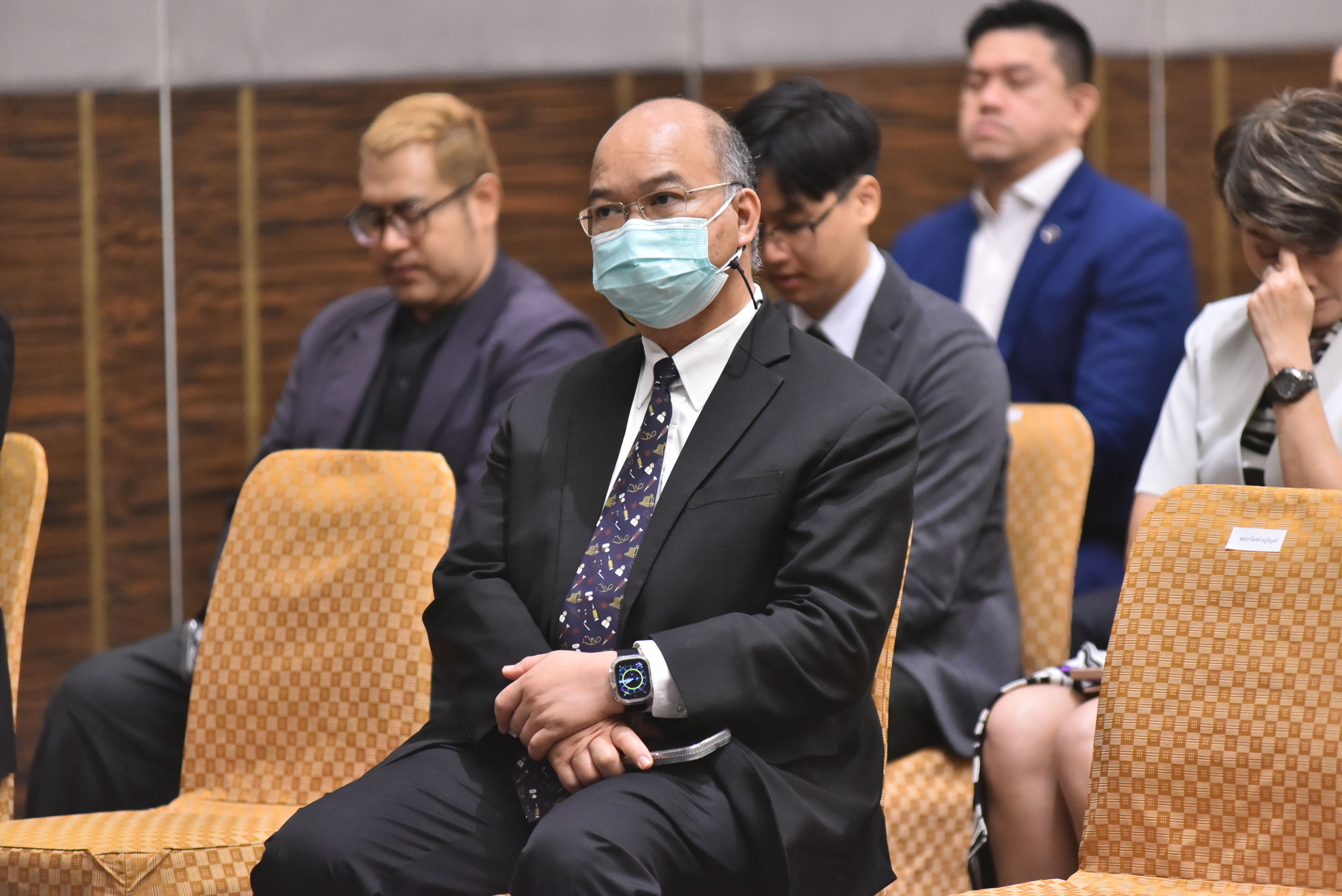 Man in dark suit and light blue face mask sits attentively at a formal event.