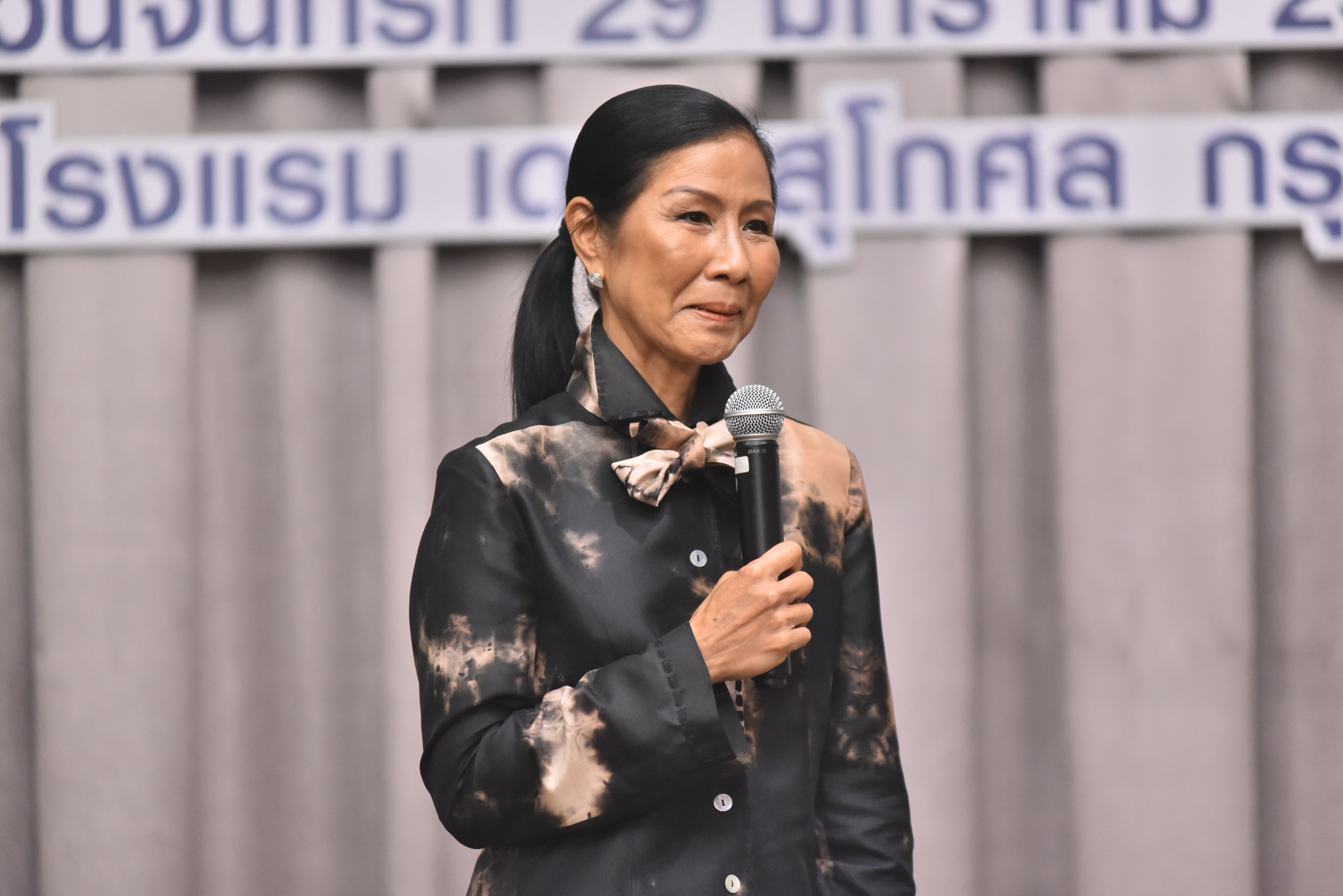 Professional woman in patterned jacket speaking into a microphone with blurred text background.