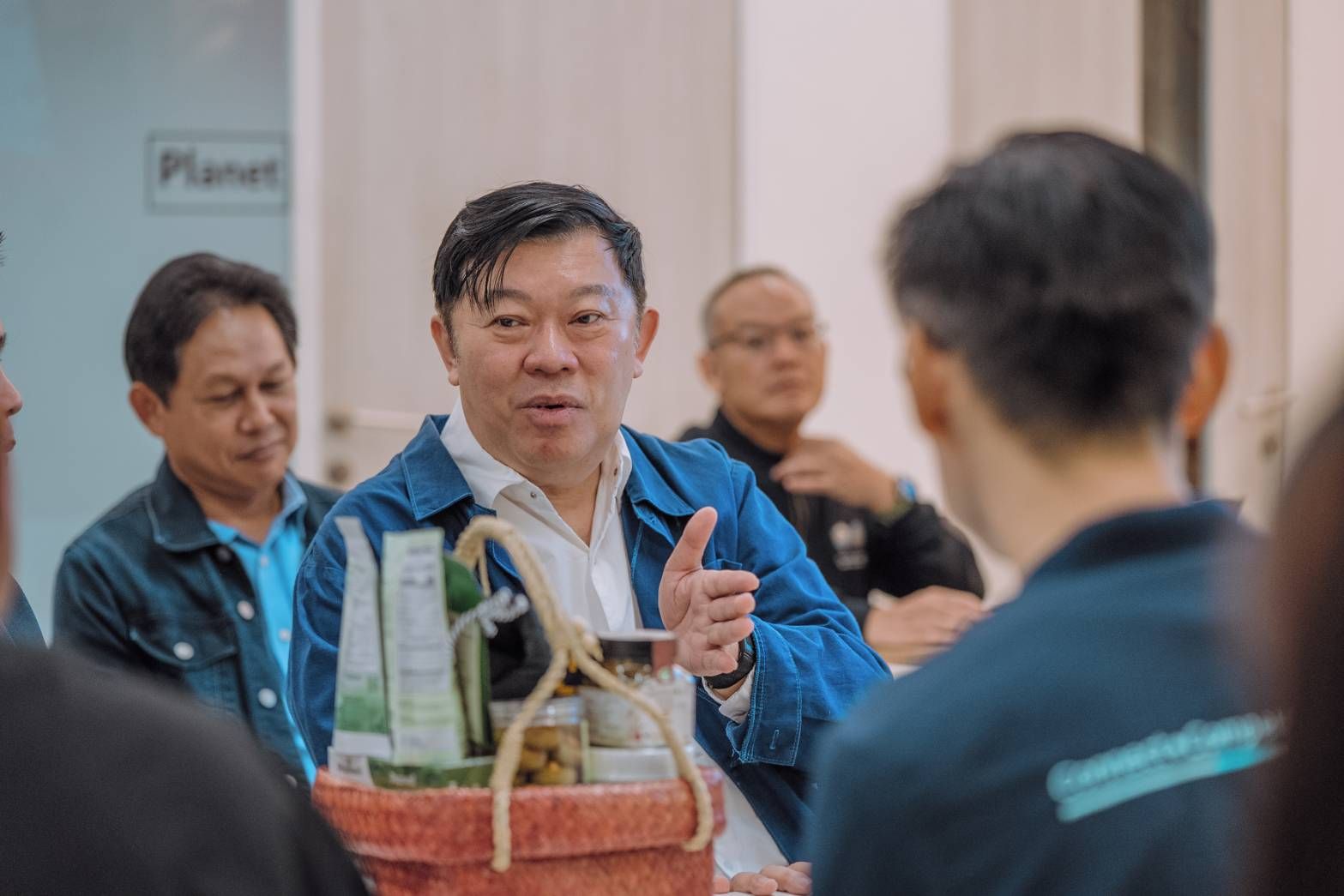 A man in a blue jacket speaks while gesturing to an attentive audience indoors.