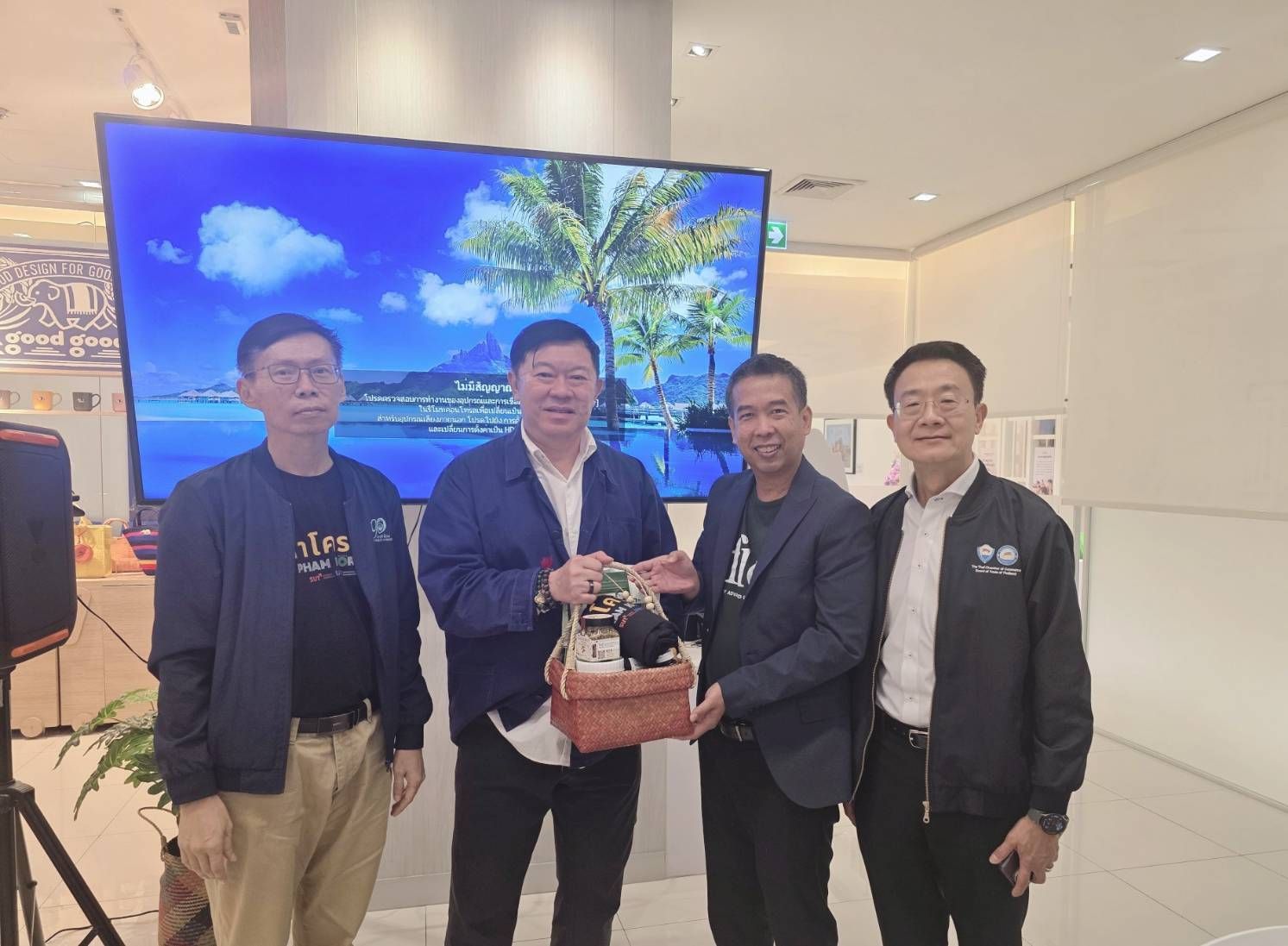 Four men smile, one holding a gift, in front of a large screen displaying a beach scene.