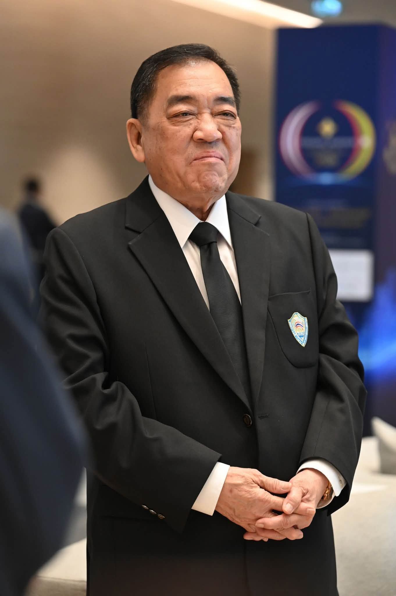 Older Asian man in black suit and tie, hands clasped, standing at a formal event.
