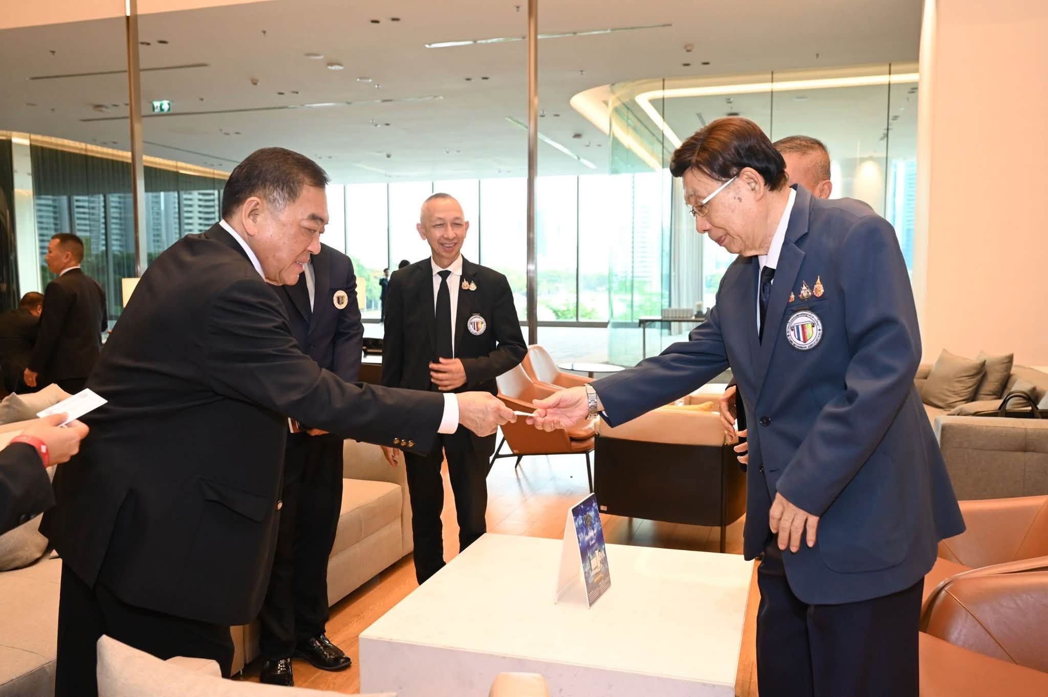 Two men in suits shake hands and exchange documents in a modern meeting room.