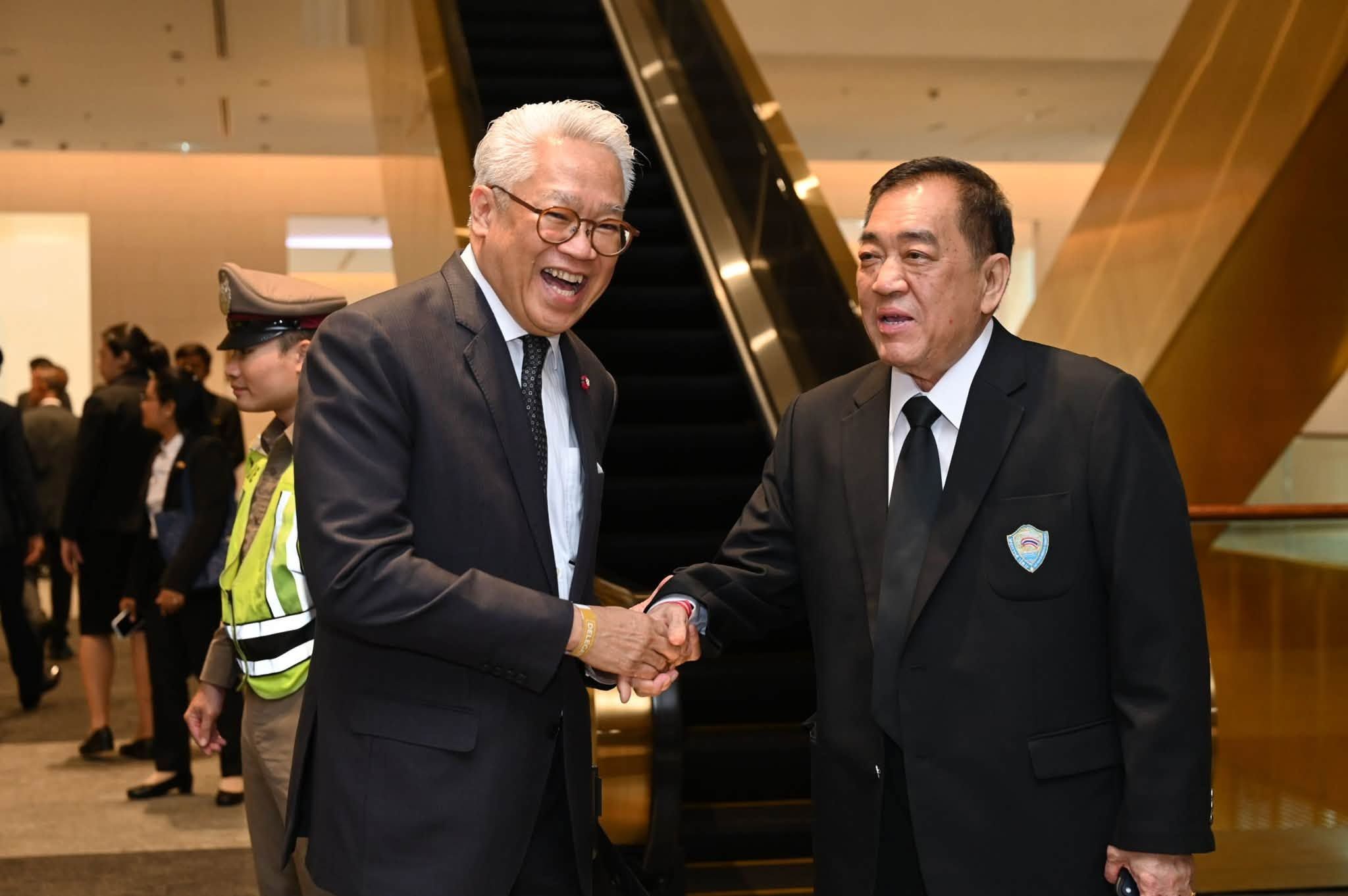 Two well-dressed Asian men shaking hands warmly in a modern building with escalators.