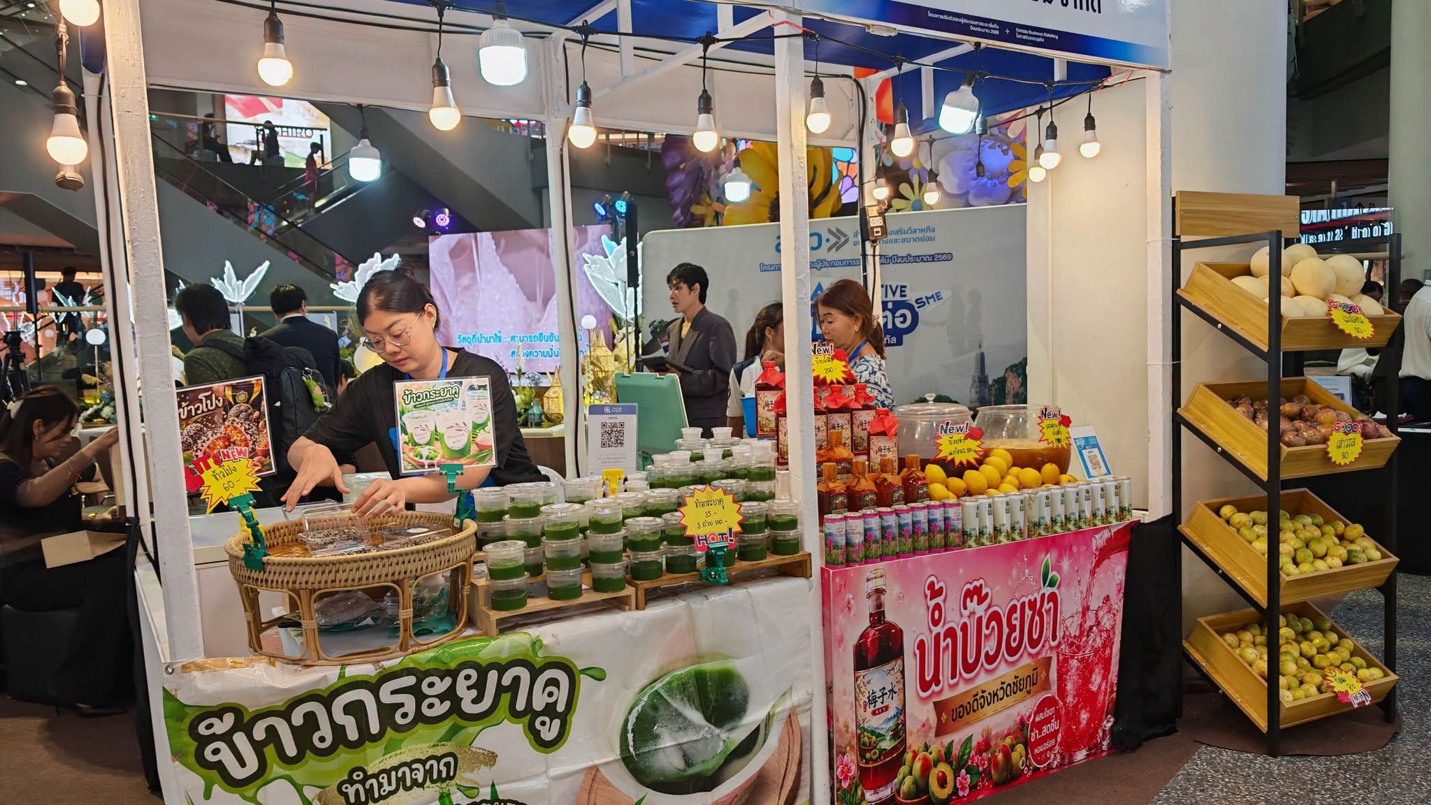 Woman serves snacks from an indoor market stall with drinks, fruits, and Thai signage.