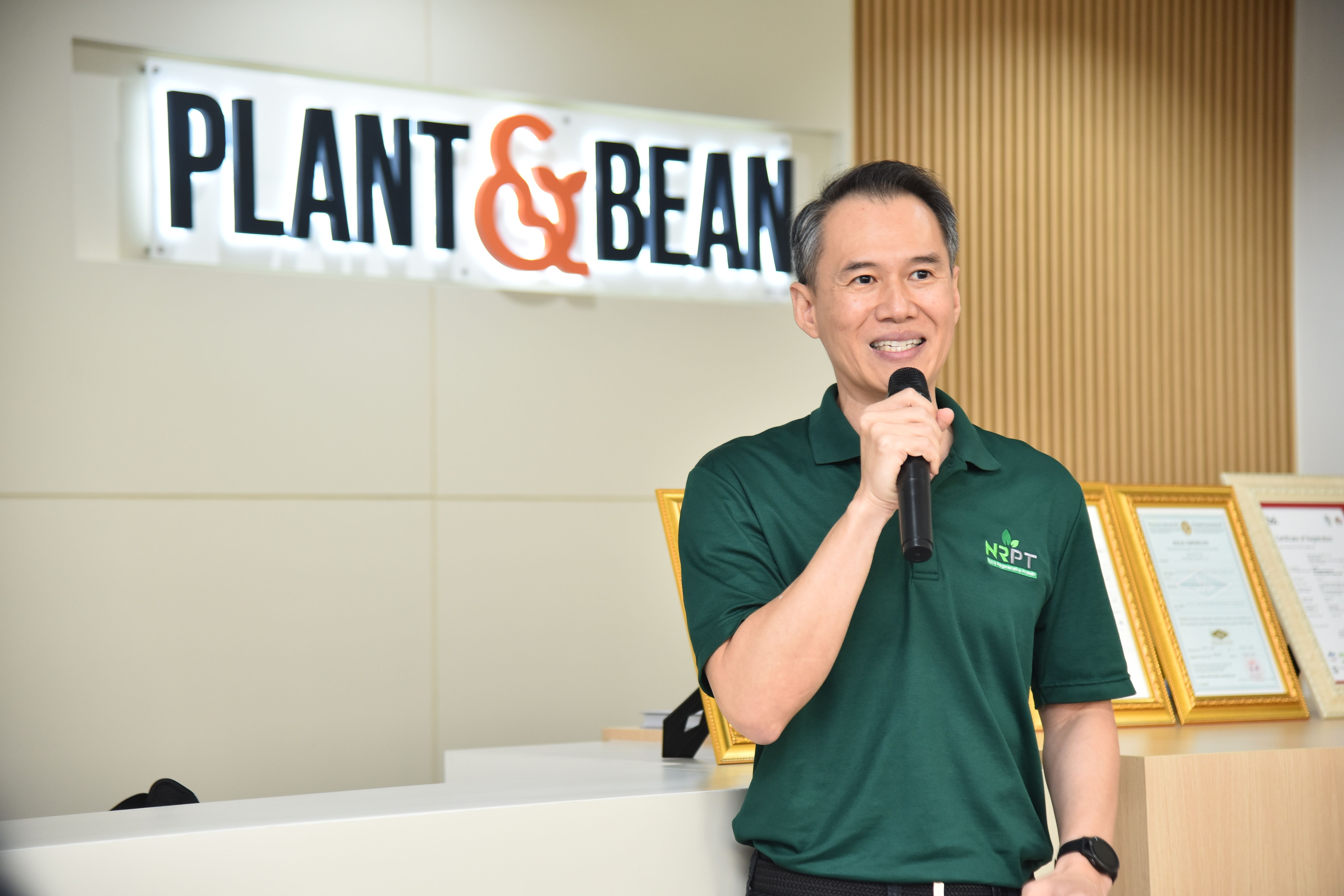 Man in green shirt speaks into microphone in front of a "PLANT & BEAN" sign.
