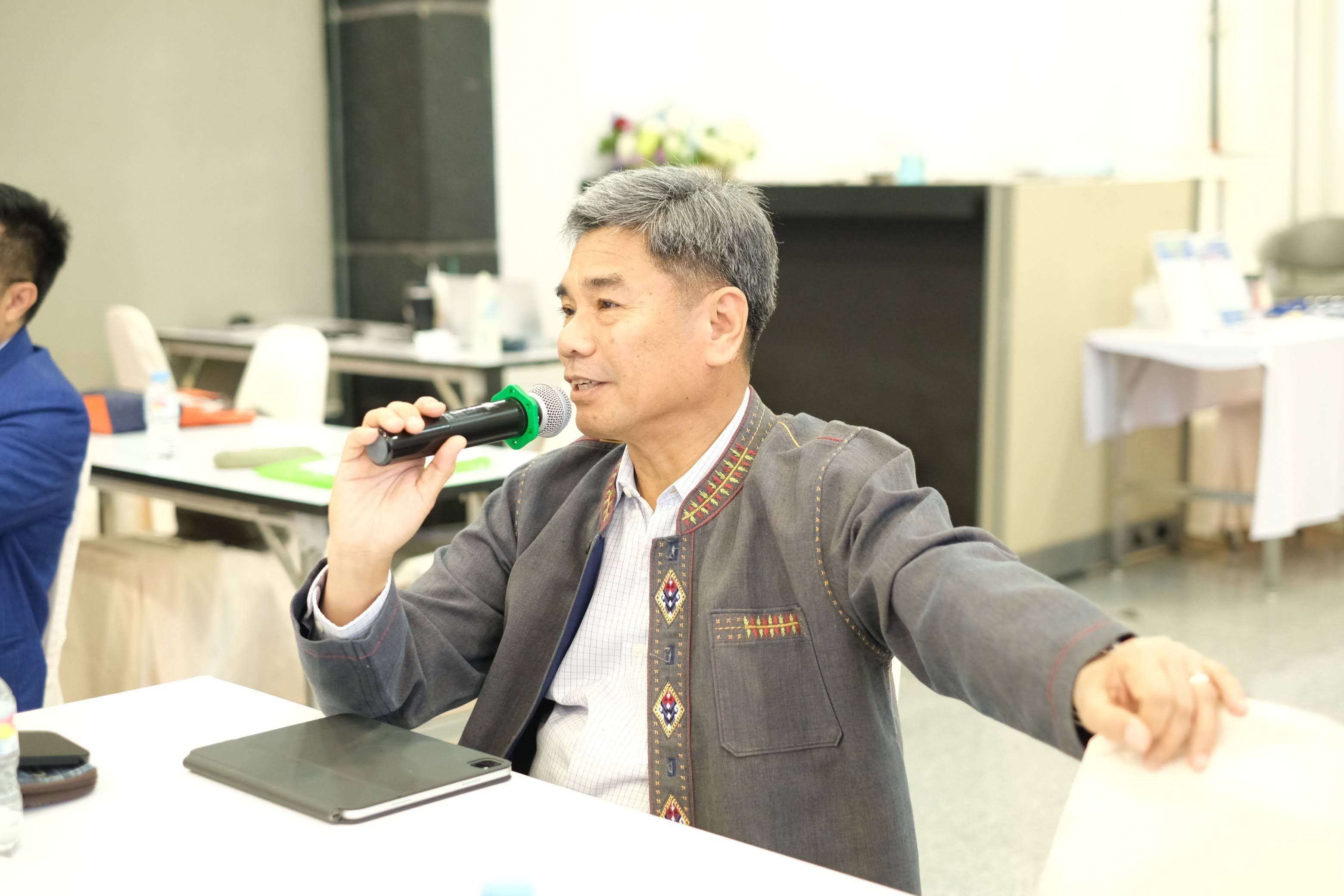 A man in a patterned jacket speaks into a microphone at a conference table.