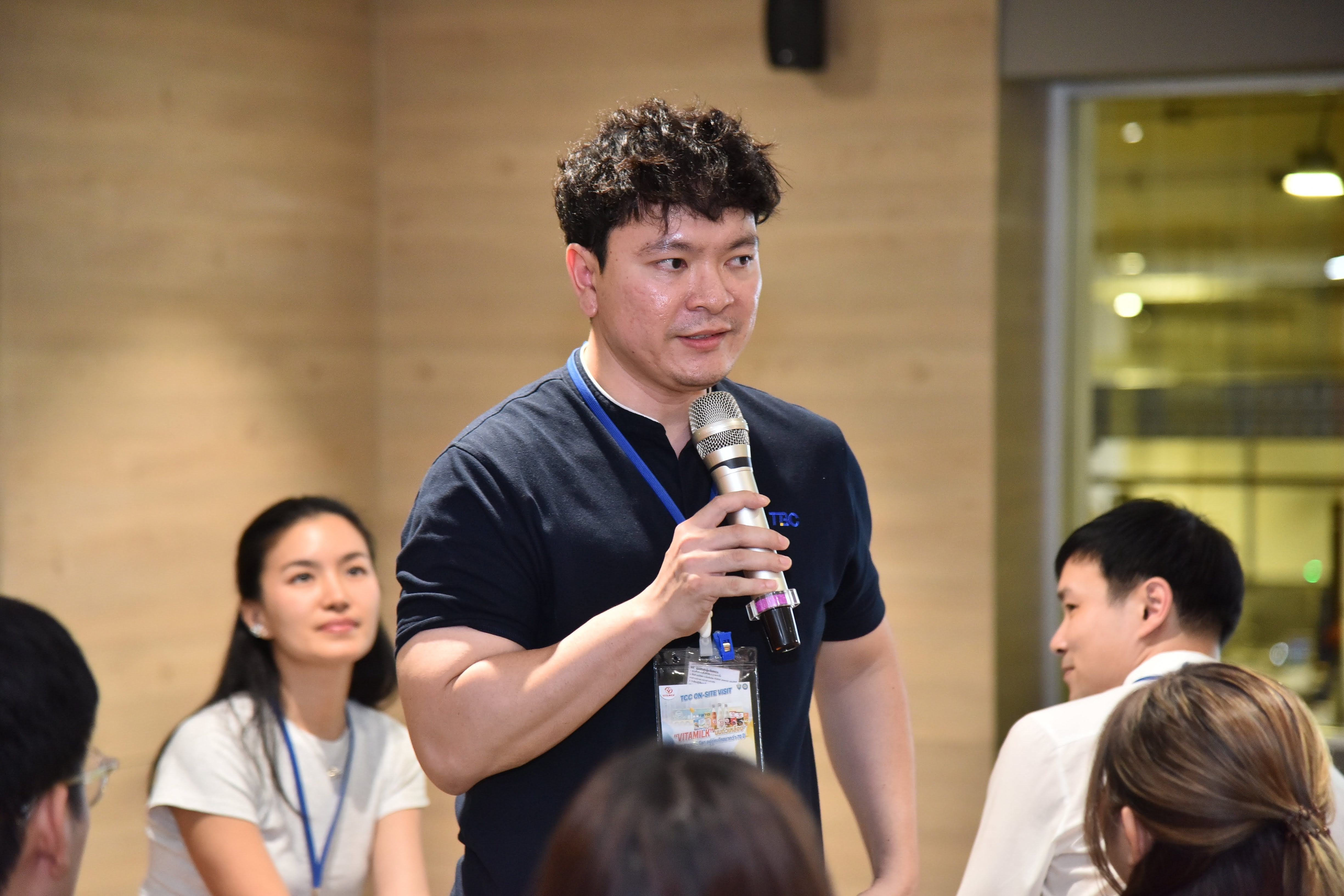 Man with curly hair in a dark shirt speaking into a microphone at an event.