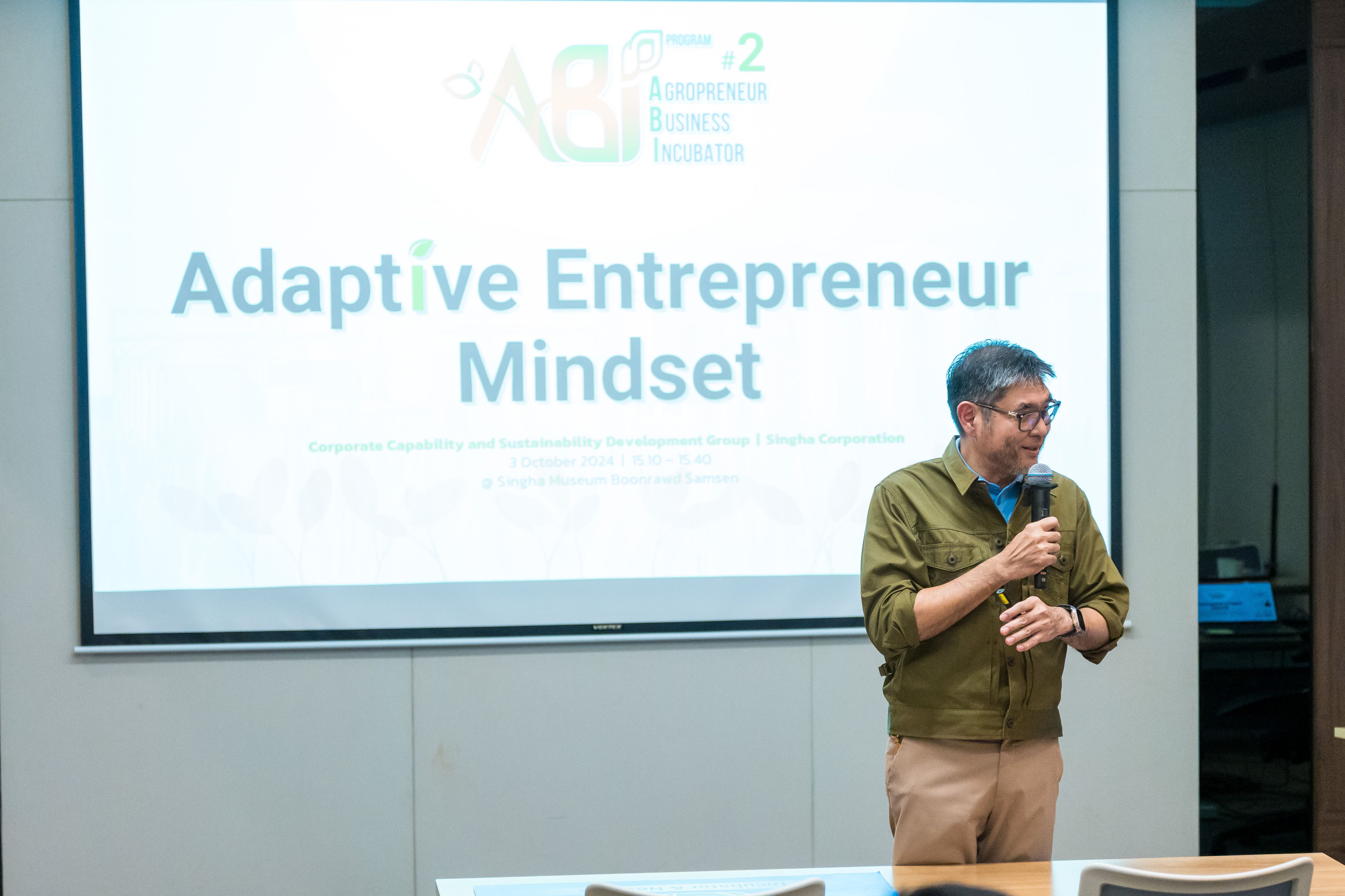 Man speaks into microphone at presentation with 'Adaptive Entrepreneur Mindset' on screen.