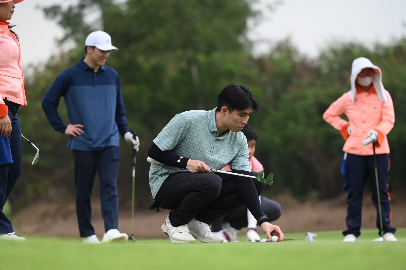 Golfer crouches to adjust a ball on a green, watched by companions.