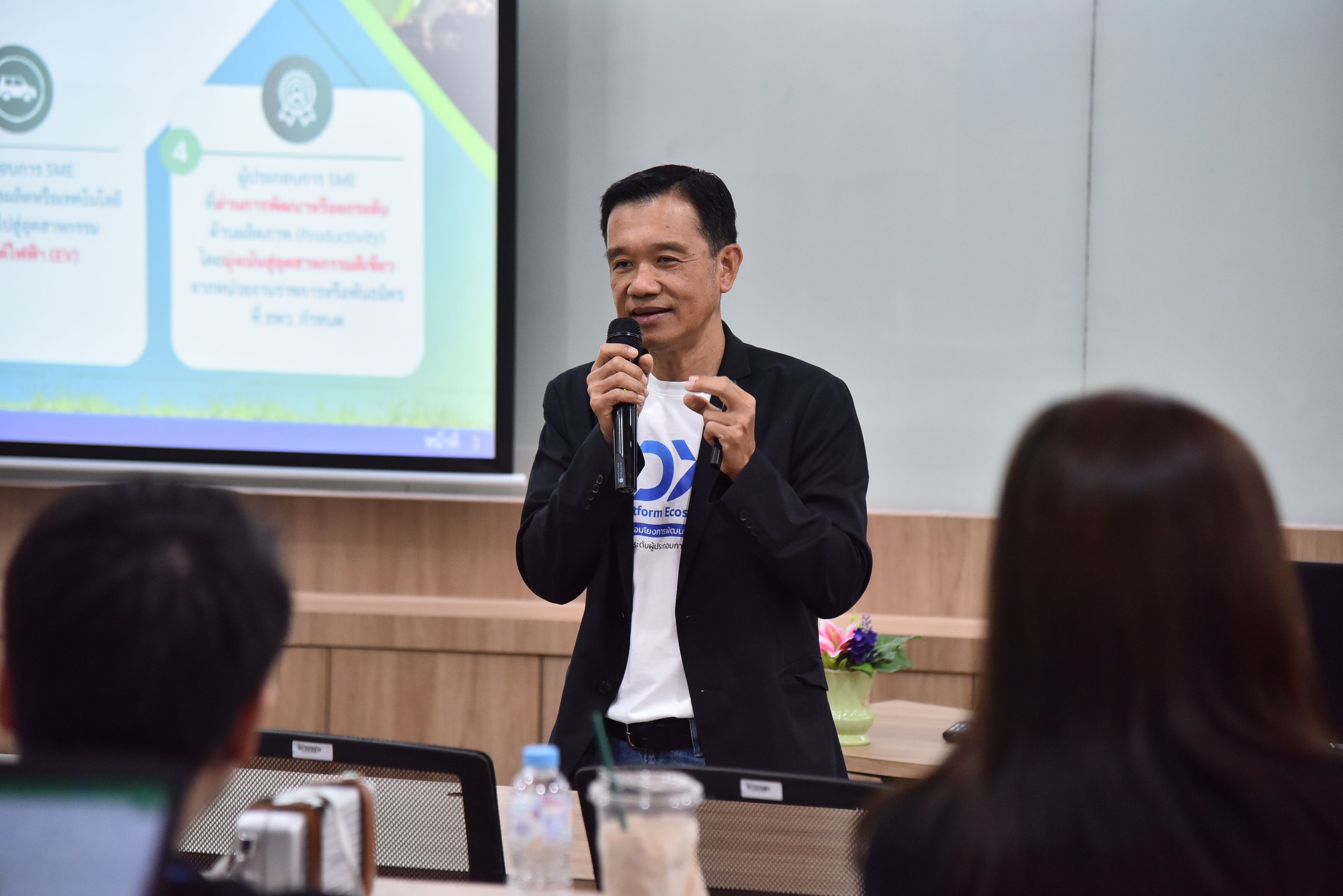 Man in black jacket speaking into a microphone during a presentation to an audience.