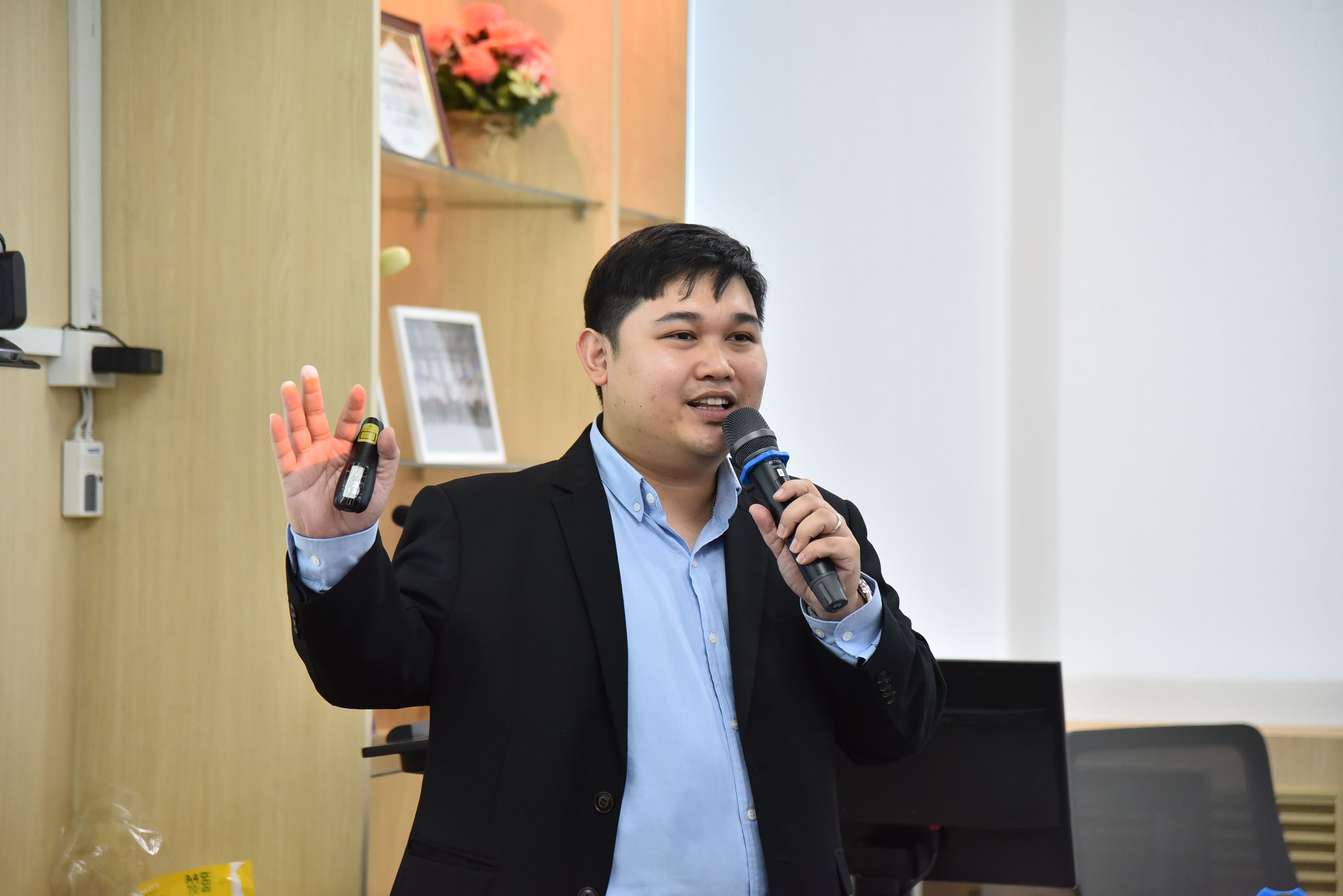 Man in black suit and blue shirt speaks into a microphone, gesturing with raised hand.