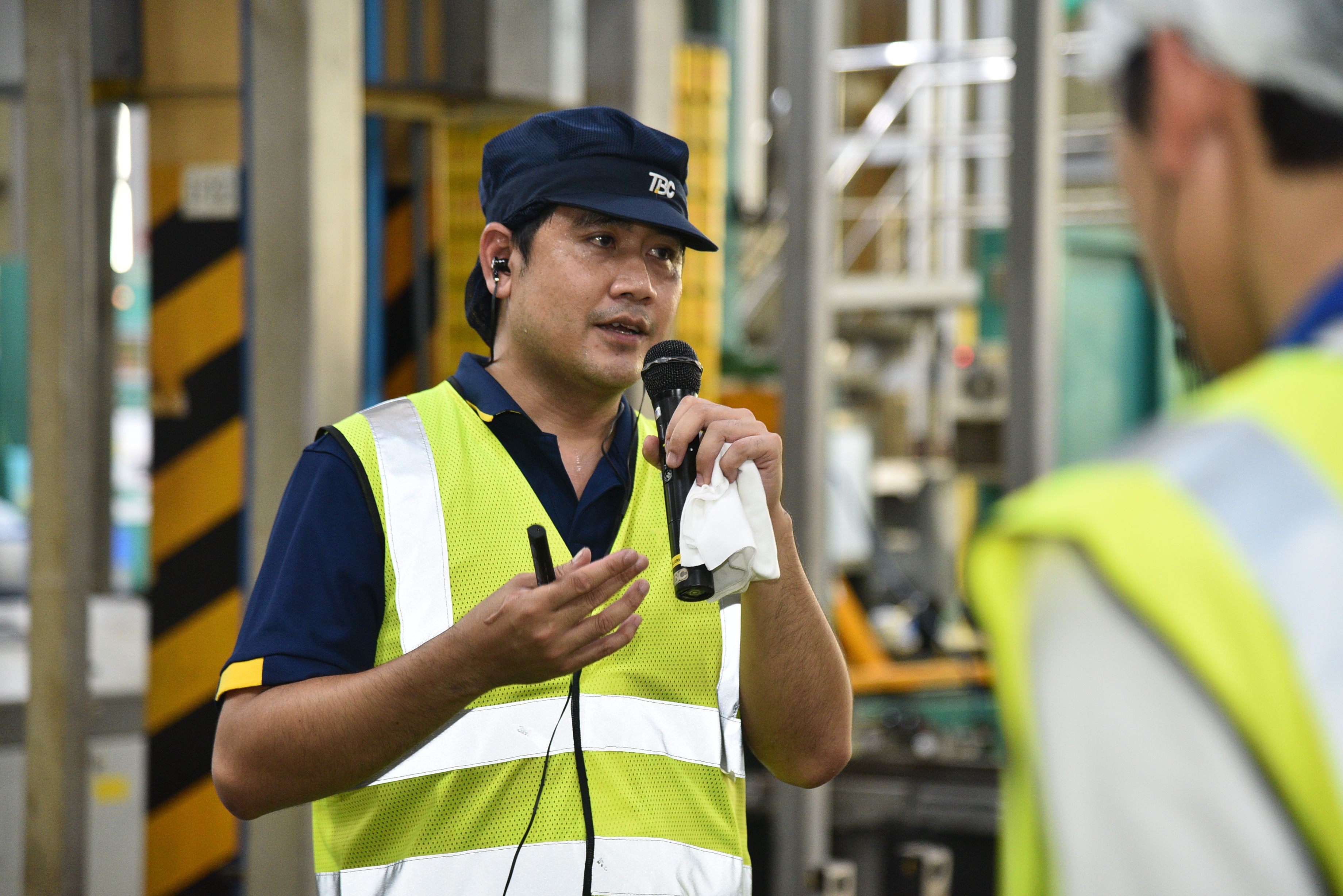 Man in safety vest and cap speaks into a microphone in an industrial setting.