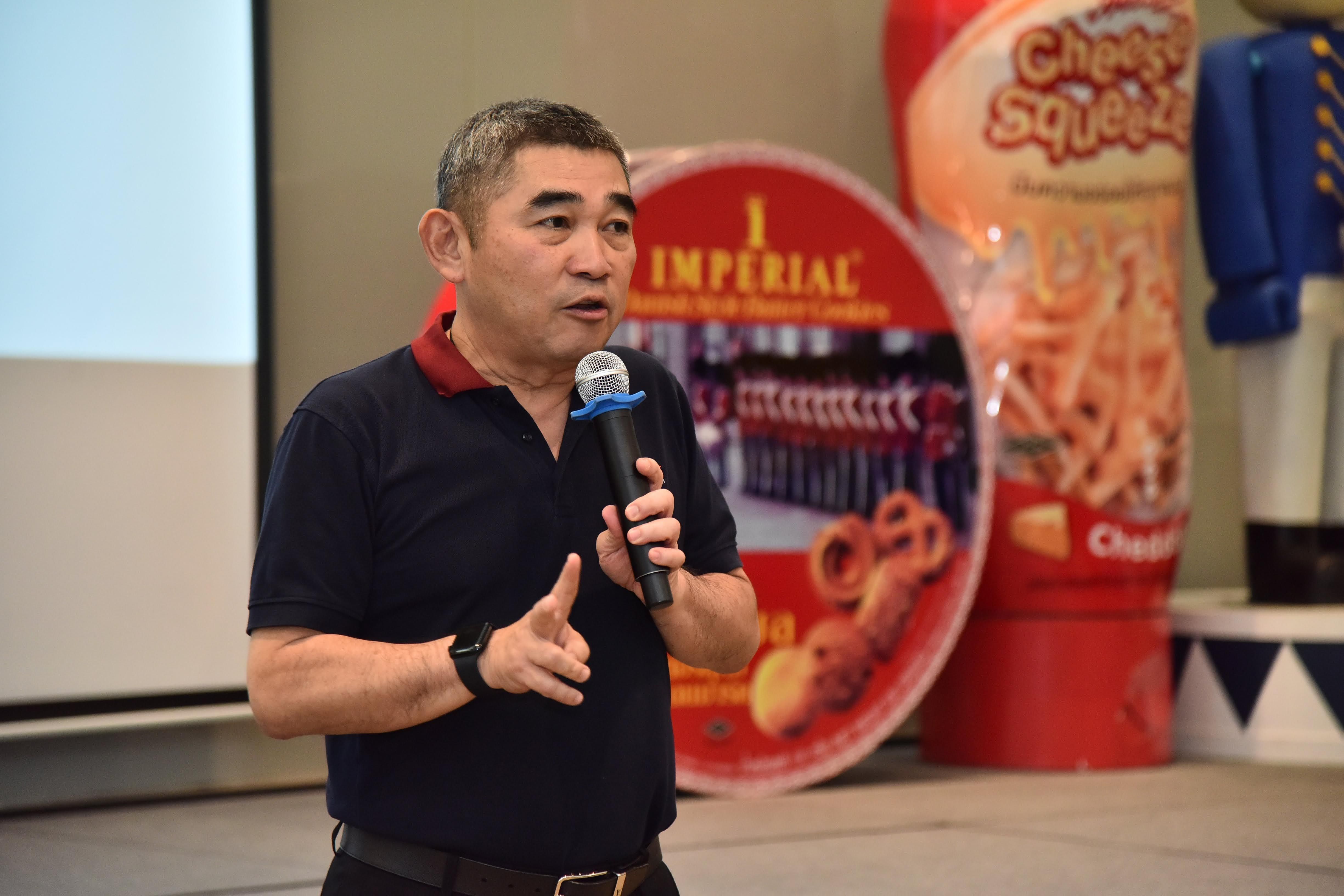 Man in dark polo shirt speaks into microphone at event with snack displays.