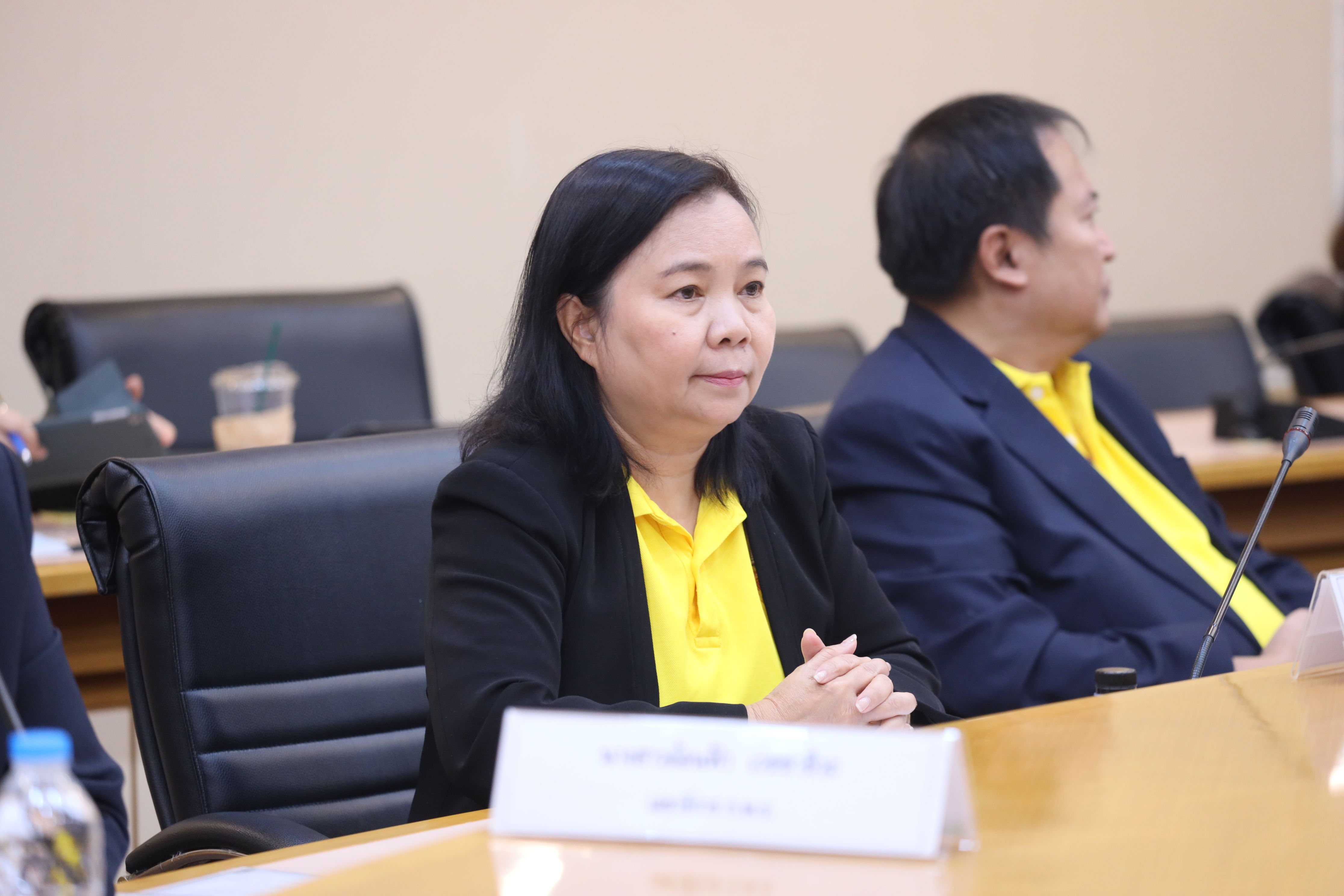 Woman in black blazer and yellow shirt at a conference table, looking attentive.