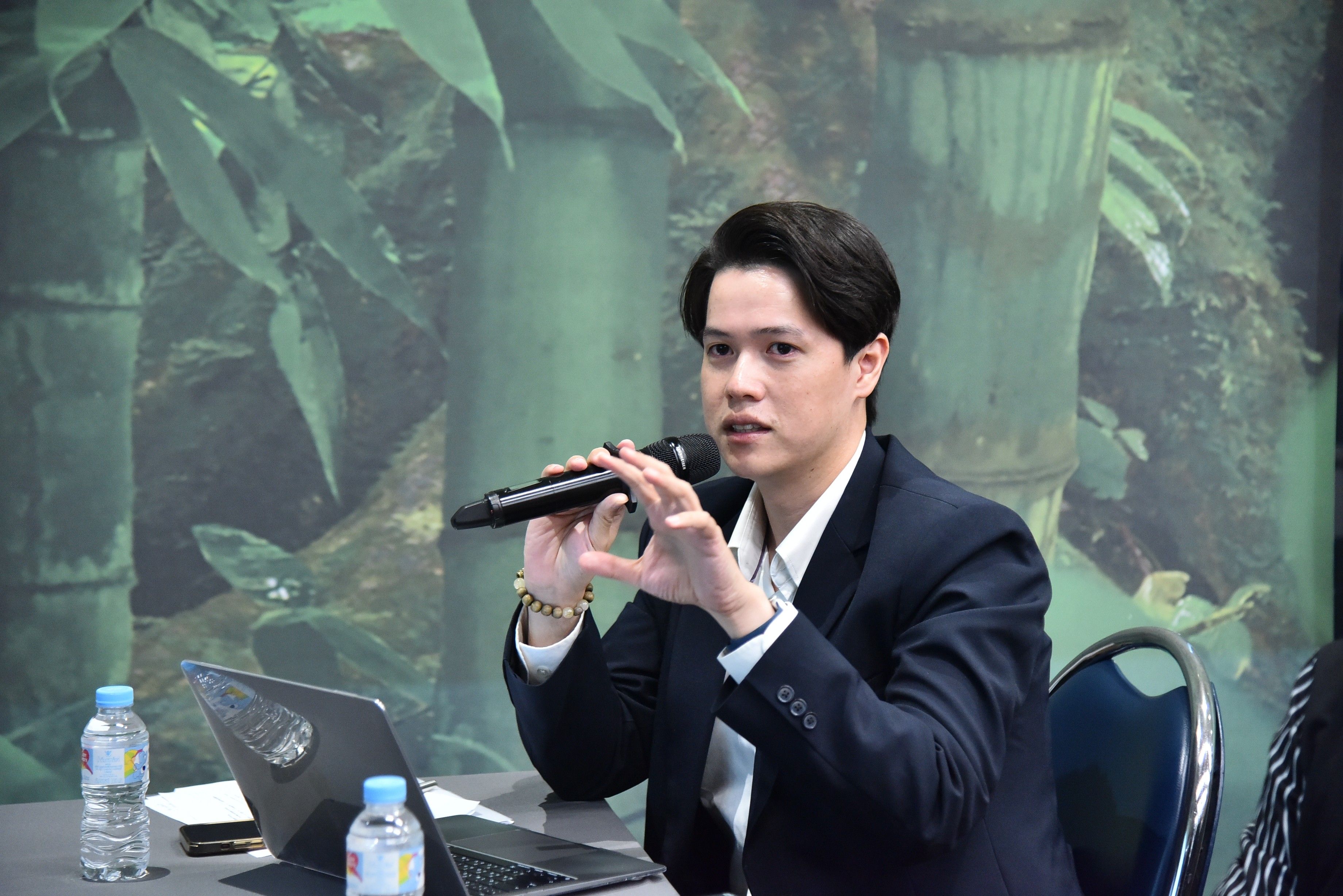 Professional man in a dark suit speaks into a microphone at a table with a laptop.